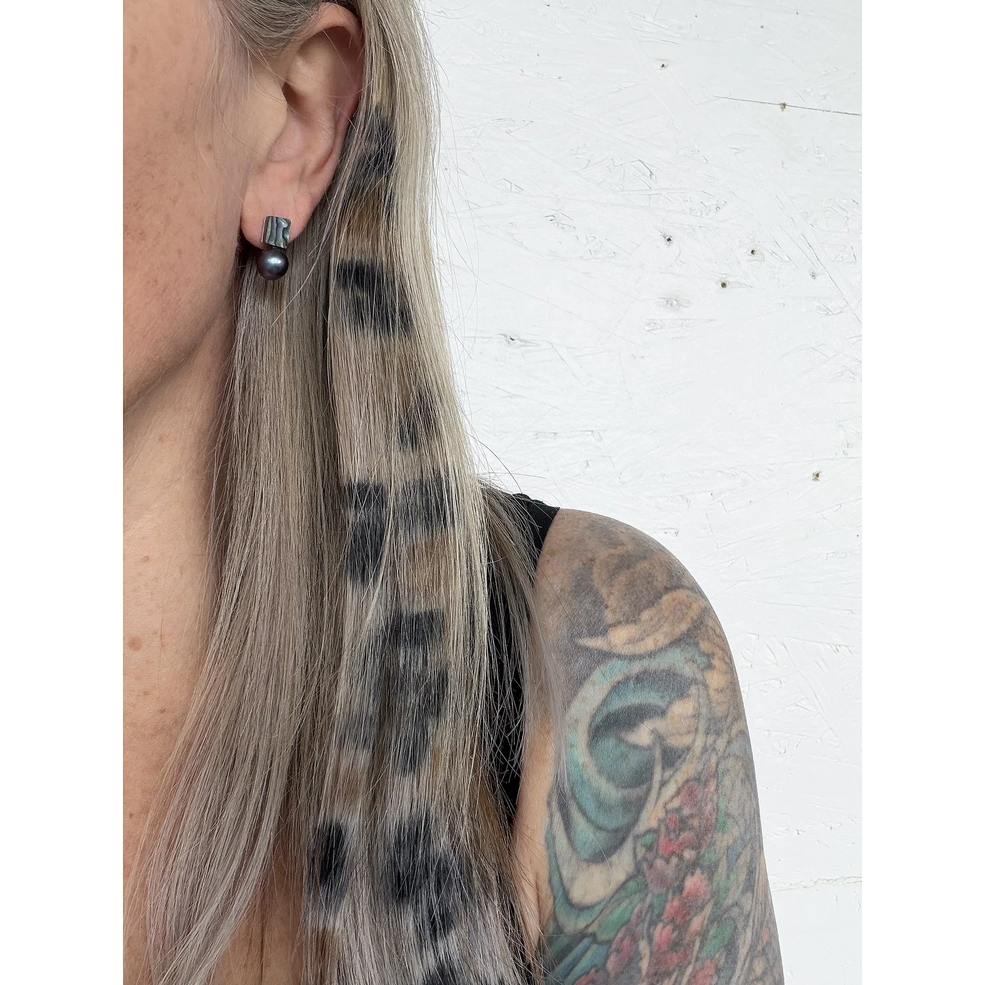 close up of woman’s ear with blonde hair and dark green abalone shell small earring and dark blue round perfect peacock pearl