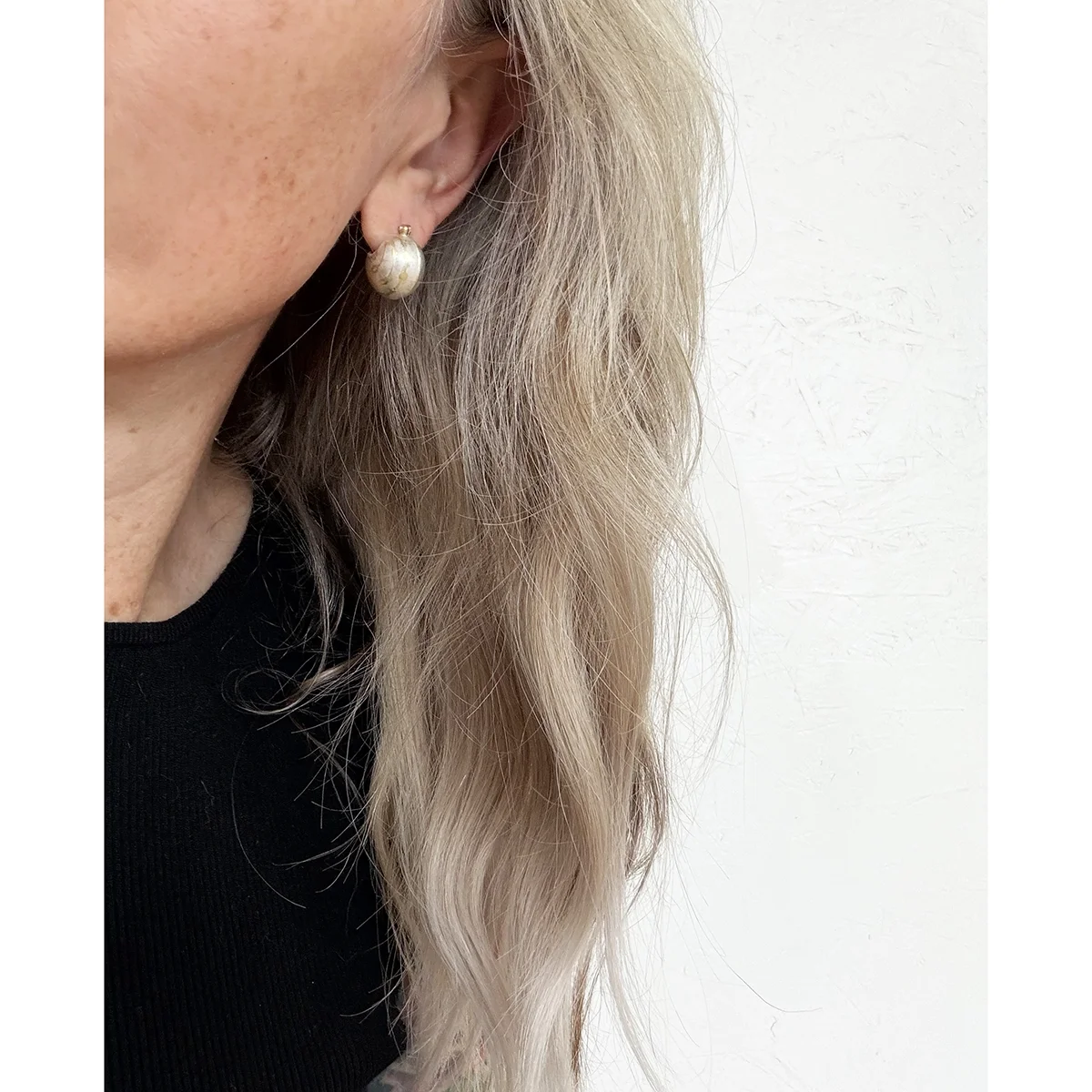 side view of woman with blond wavy hair and black shirt wearing small silver ball earring with gold lines