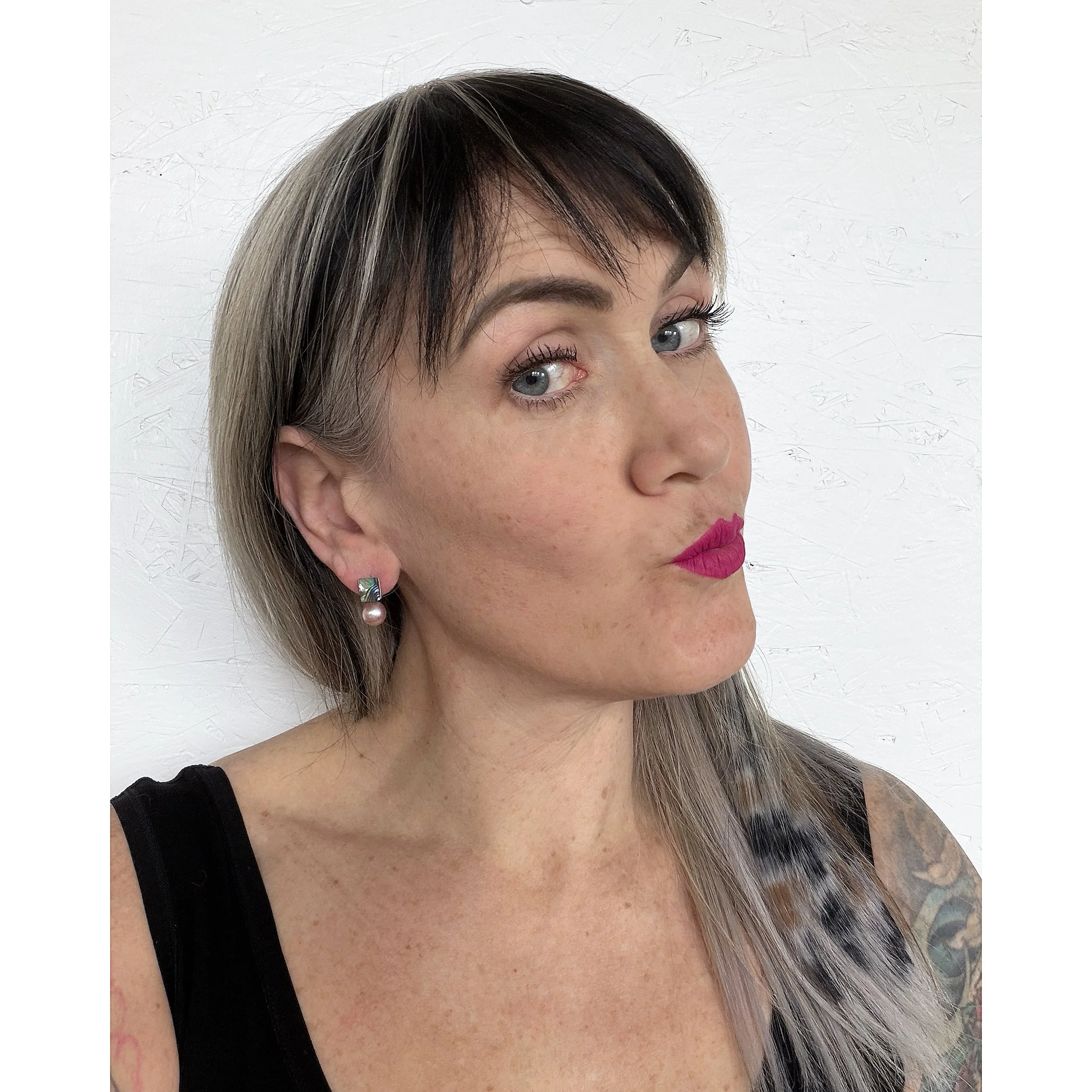 woman with blue eyes blonde hair and pink lipstick wearing small earring with waves of blue, green, pink and blue on a small shell piece next to a big pink freshwater pearl