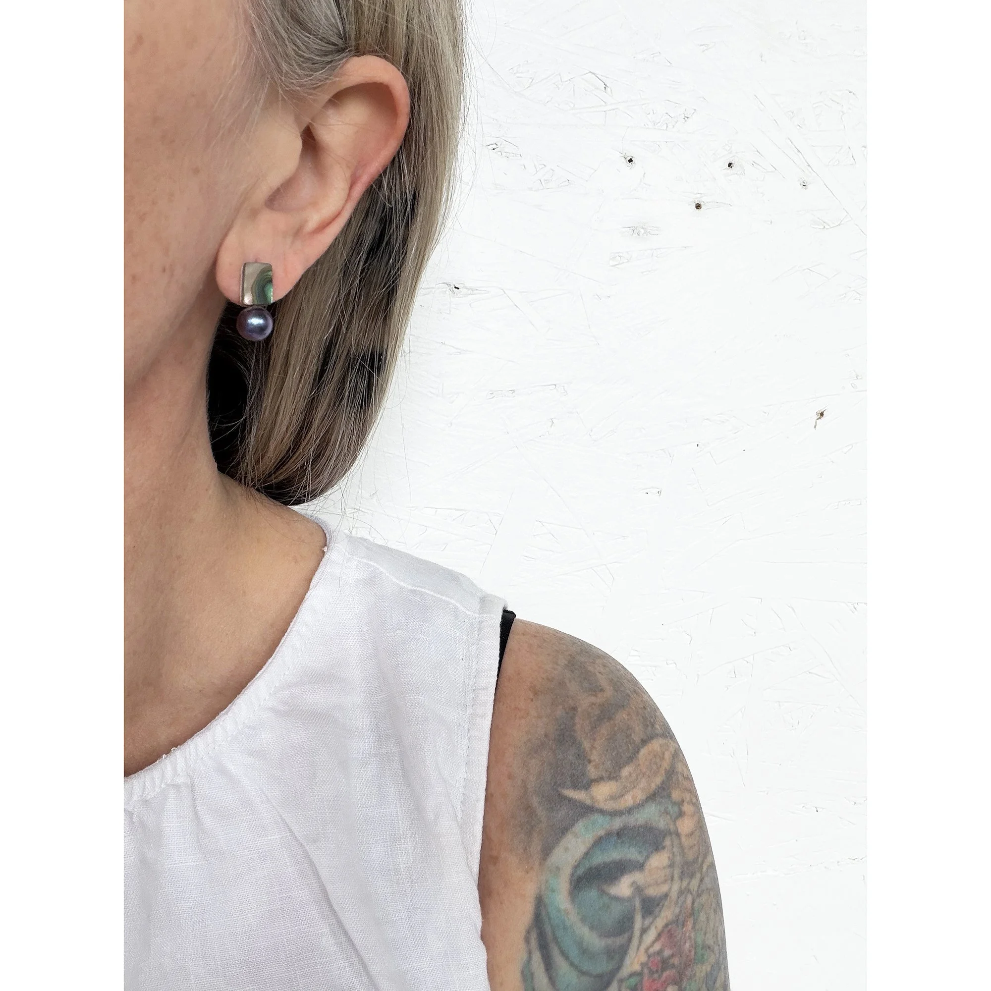 side view of woman with blonde hair and white shirt wearing small pearl stud earring with a rainbow of color on top and a round black pearl on the bottom