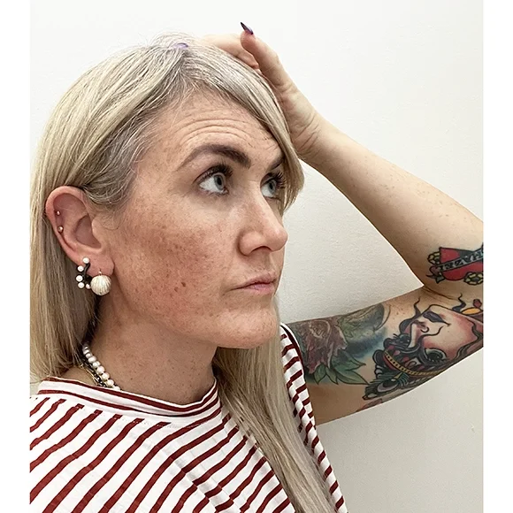 woman with blond straight hair and freckles, a red and white striped shirt and tattoos on her arms wearing small silver ball earrings and small pearl hoop earrings