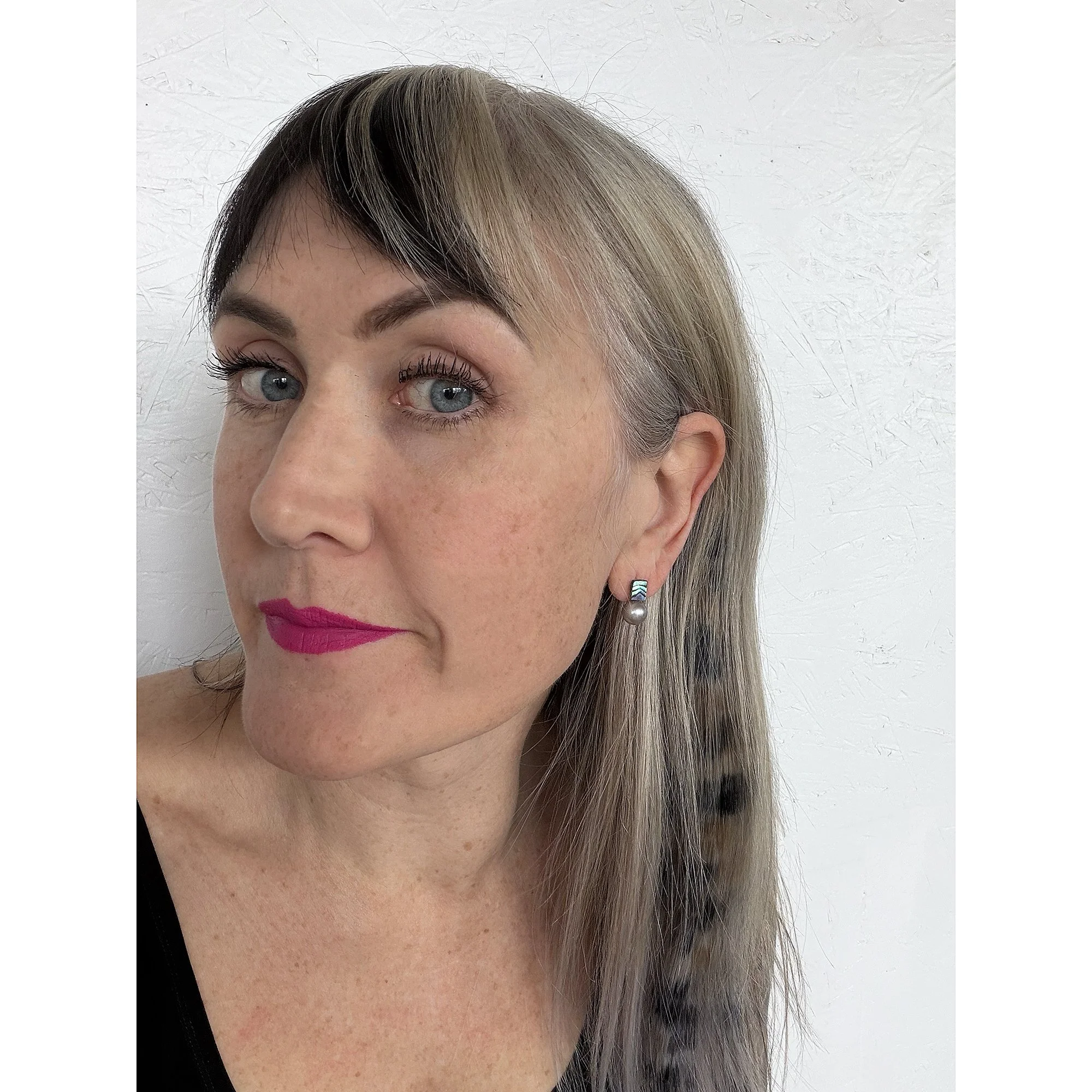 front view of woman with blonde hair, pink lipstick and white shirt wearing small pearl stud earrings with a rainbow of color on top and a round black pearl on the bottom
