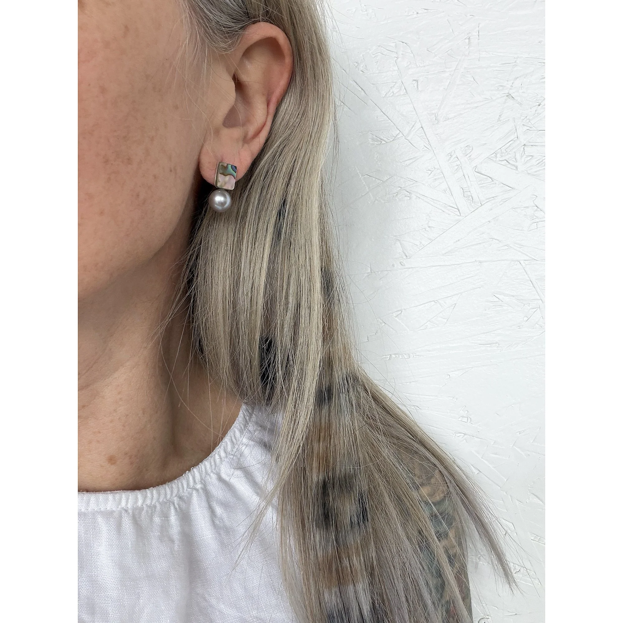 side view of woman with blonde and cheetah patterned hair and white shirt wearing small pearl stud earring with waves of color on top and a round silver pearl on the bottom