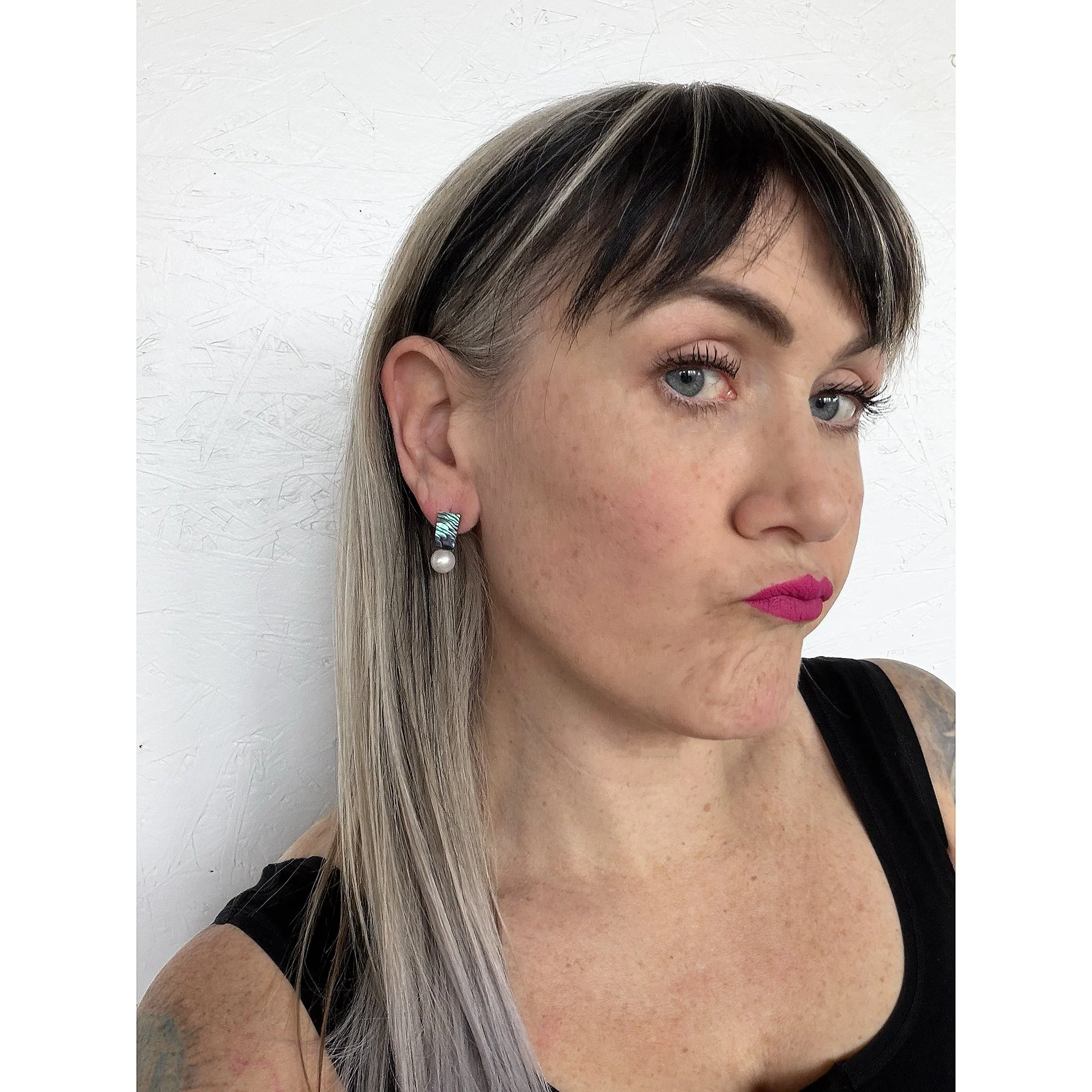front view of woman with blonde hair, pink lipstick and black shirt wearing small dark green abalone and shimmery white freshwater pearl earrings looking at camera