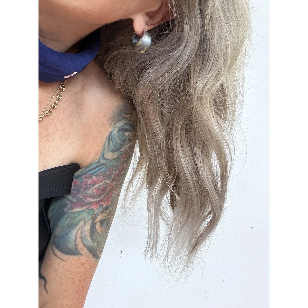 oxidized sterling silver curvy hoop earring with gold lines on woman with wavy blond hair a black sleeveless top, tattoos on her arms and a dark navy scarf on her neck