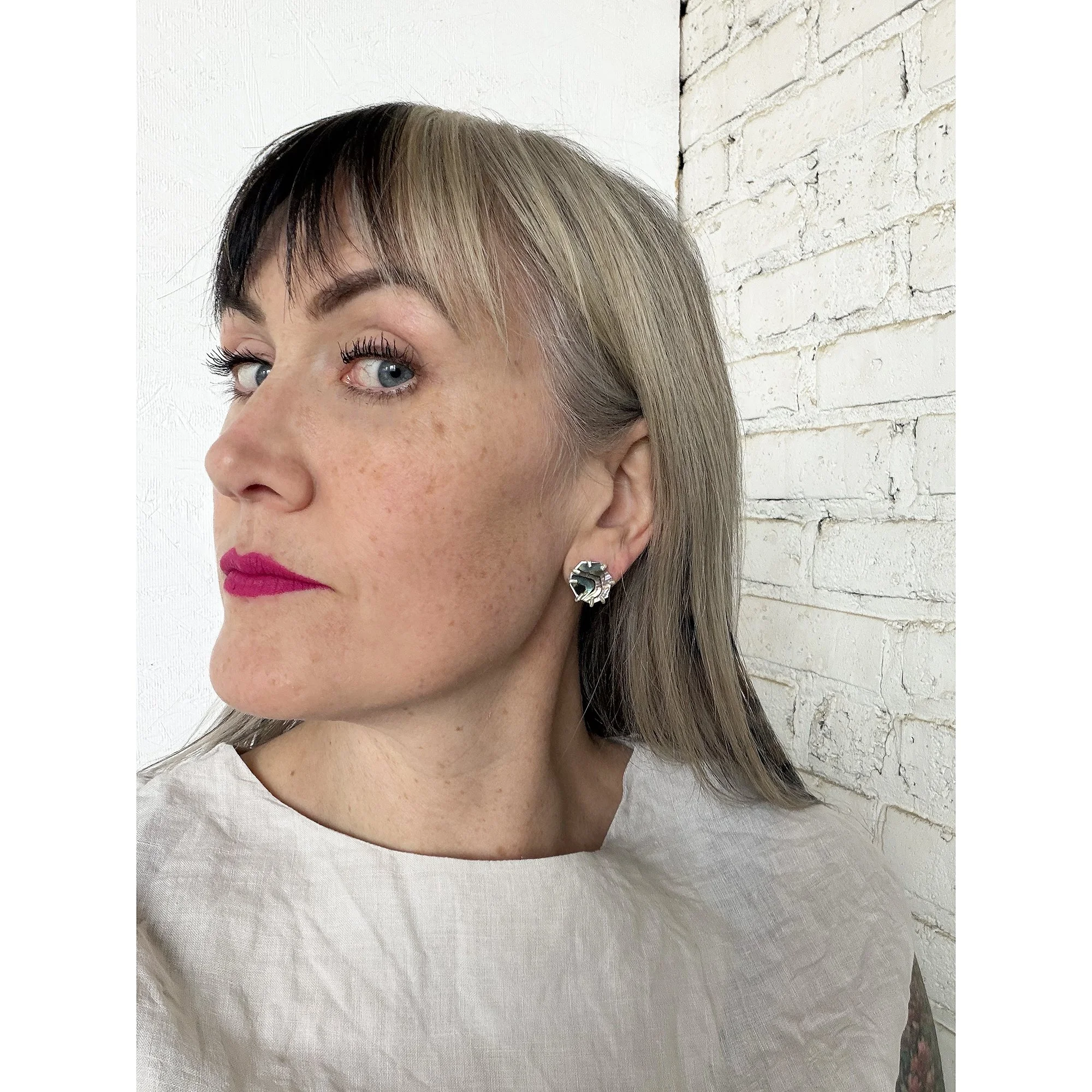 small post abalone earrings on white woman with pink lips in room with white brick