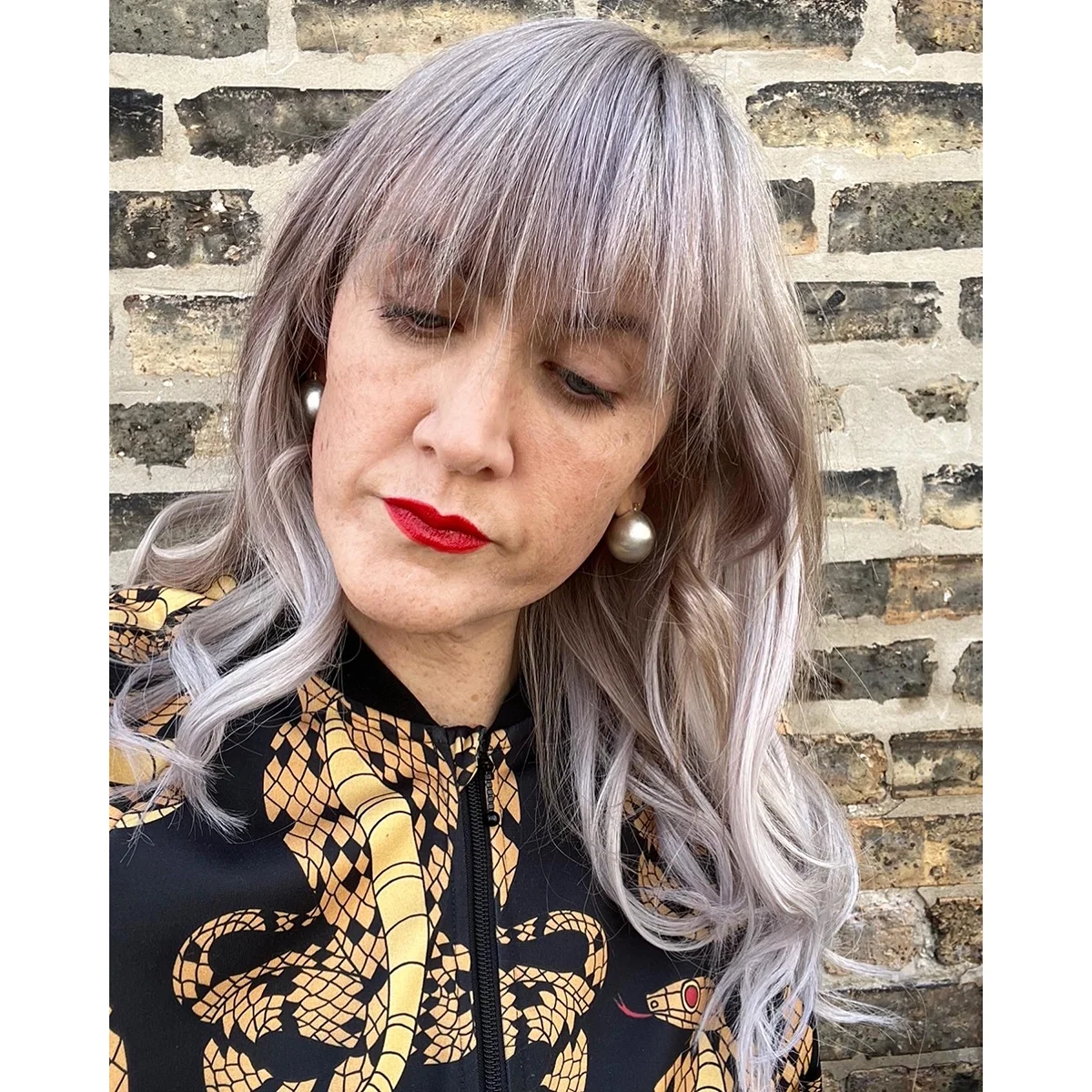 woman with blond and purple curly hair wearing a snake jacket in front of a brick wall wearing shiny silver ball earring