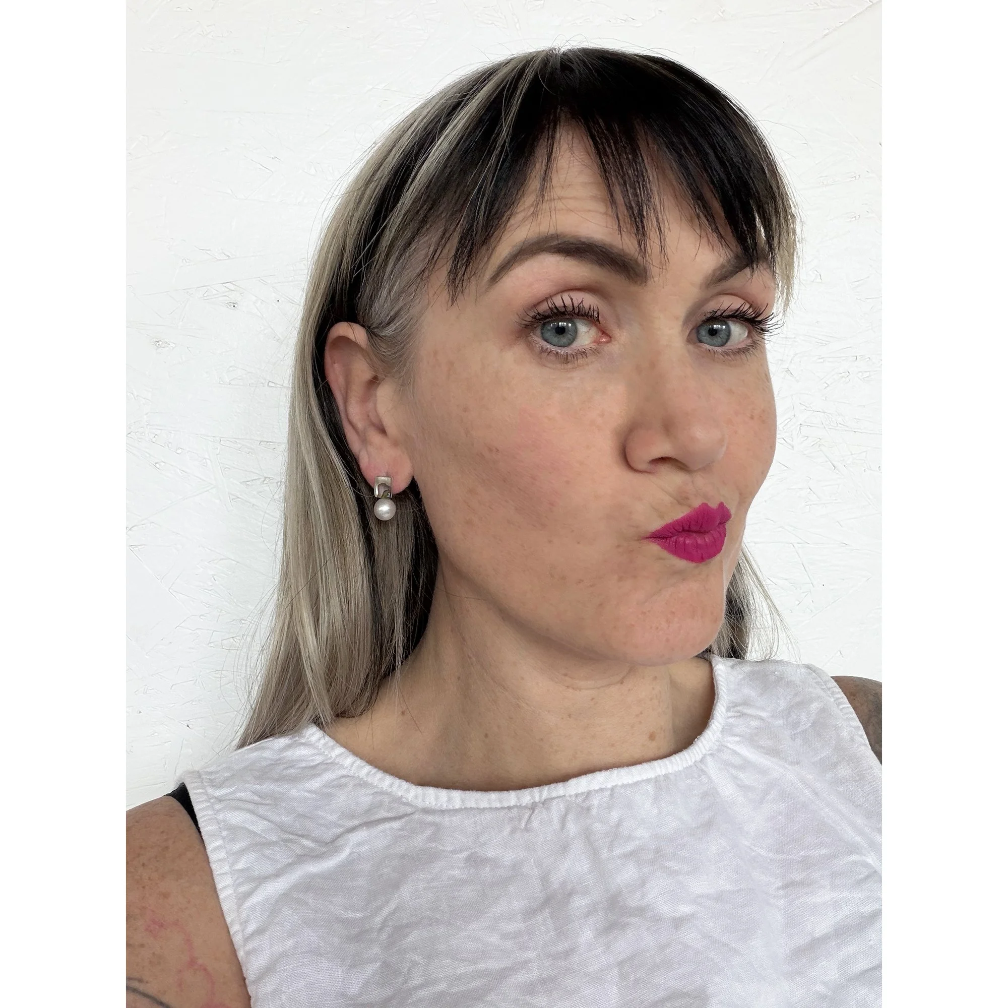 front view of woman with blonde hair and pink lipstick making kiss face wearing small pearl and abalone earring