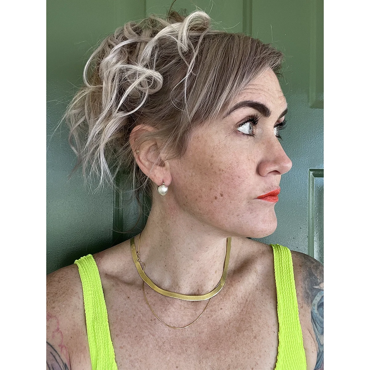 woman with blond wavy hair freckles, a neon yellow top and red lips wearing small shiny sterling silver ball earrings in front of a dark green door
