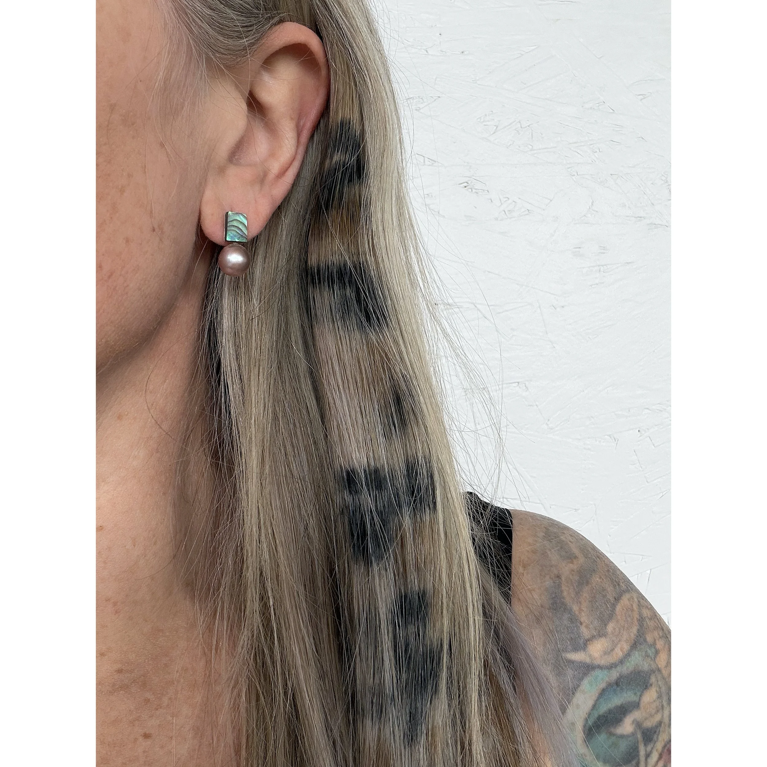 close up of woman’s ear with blonde hair and big pink freshwater pearl next to small abalone shell piece with waves of blue, green, pink and blue