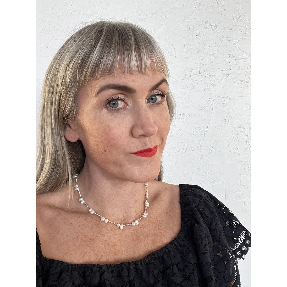 woman with blonde hair and red lips wearing dark blue lace off shoulder top and delicate single strand white and silver freshwater pearl necklace