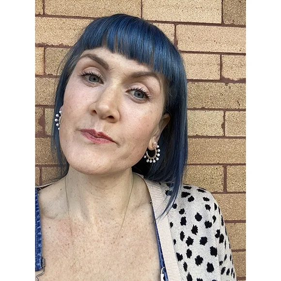 woman with blue hair facing camera wearing silver hoop earrings with white pearls in front of brick building