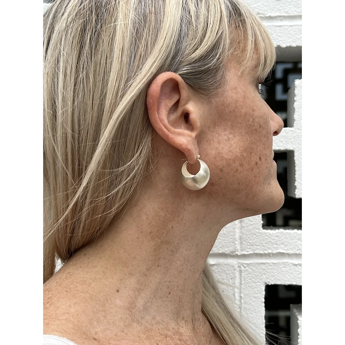 woman with straight blond hair and bangs wearing sterling silver bauble hoop earrings in front of white and black bricks