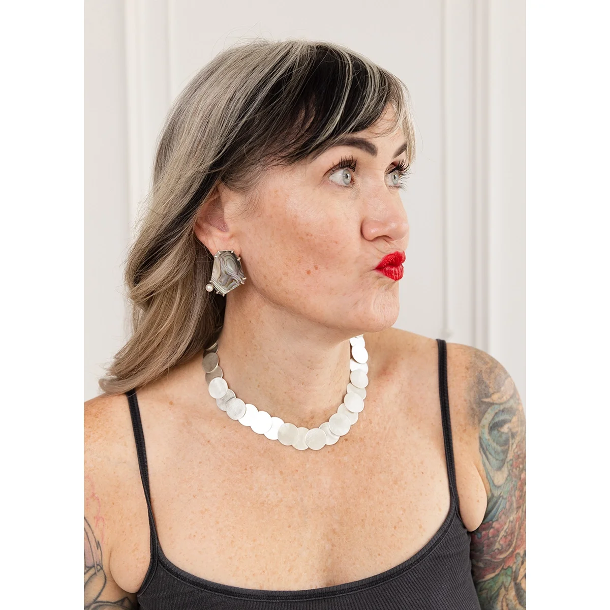 woman with black and white hair and shiny silver necklace wearing pink green and purple shell earrings with white round pearl