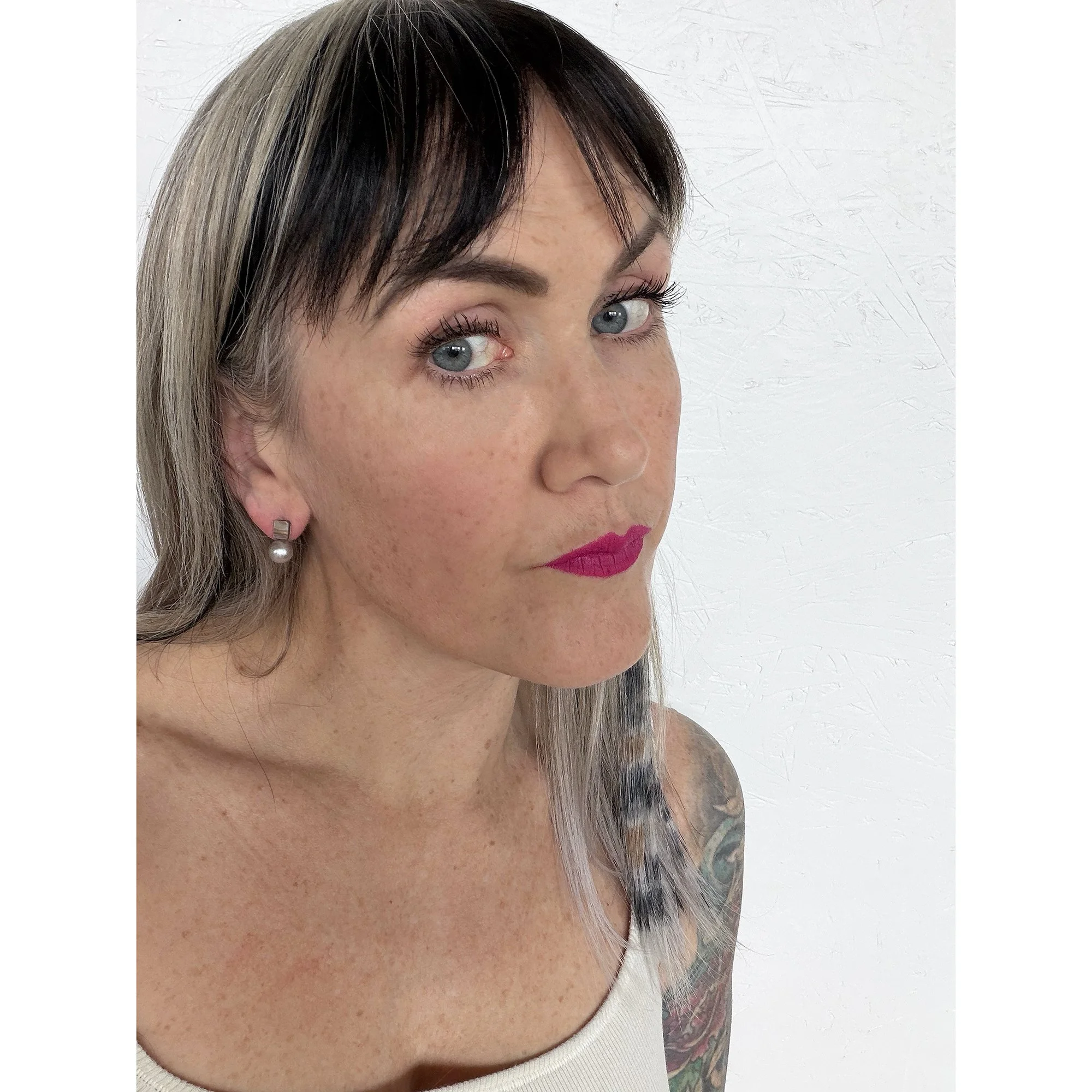 front view of woman with blonde hair, pink lipstick and white shirt wearing small pearl stud earrings with a rainbow of color on top and a round silver pearl on the bottom