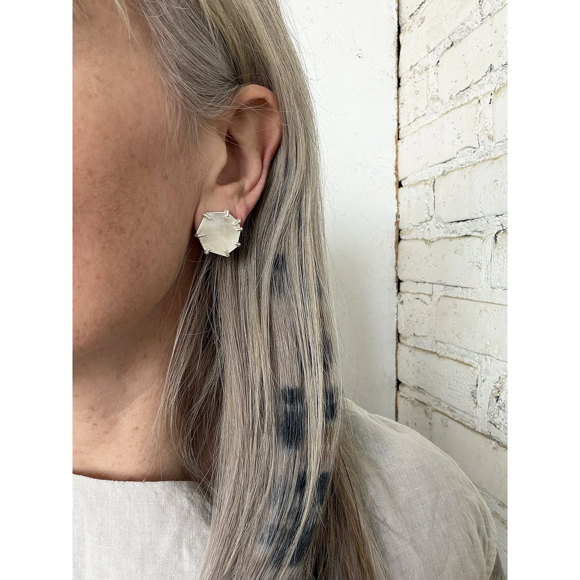 close up of mother of pearl post earring on woman with blonde hair in room with white brick