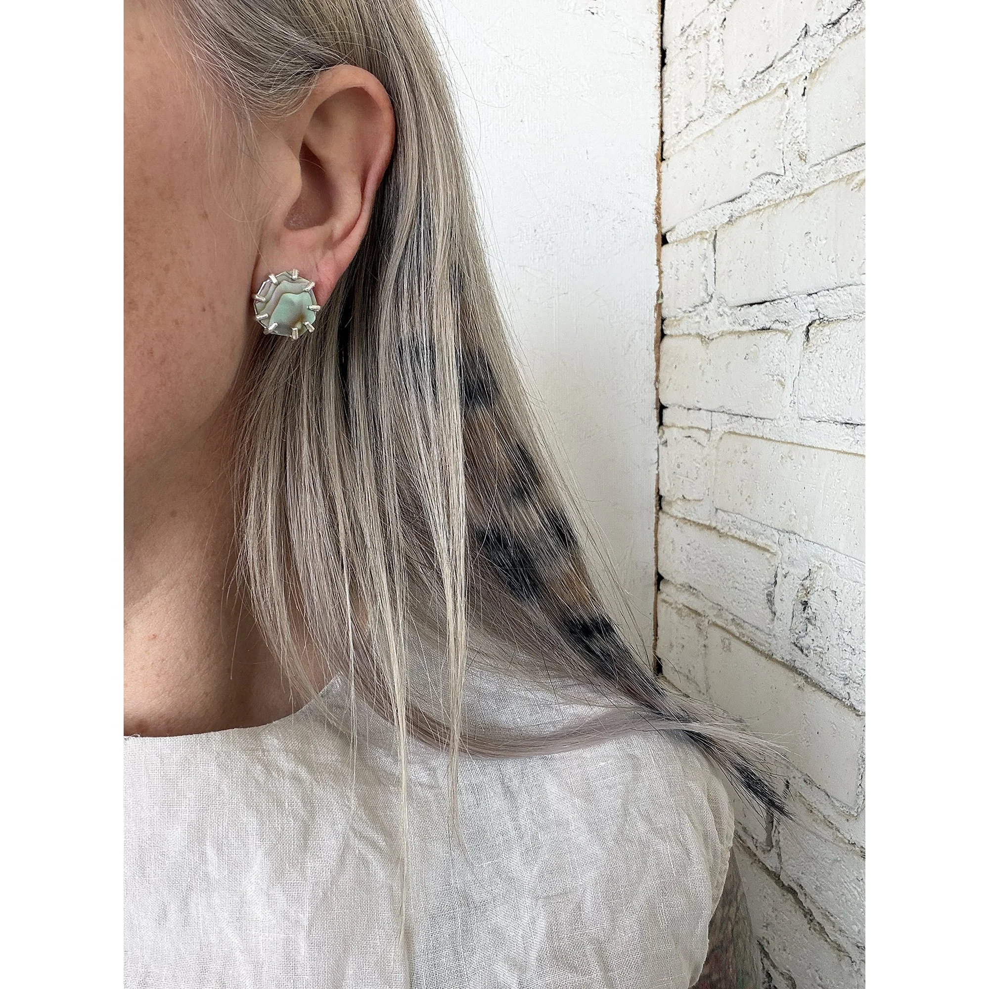blonde woman's ear with shimmery small red and pink abalone post and silver prongs with white brick background