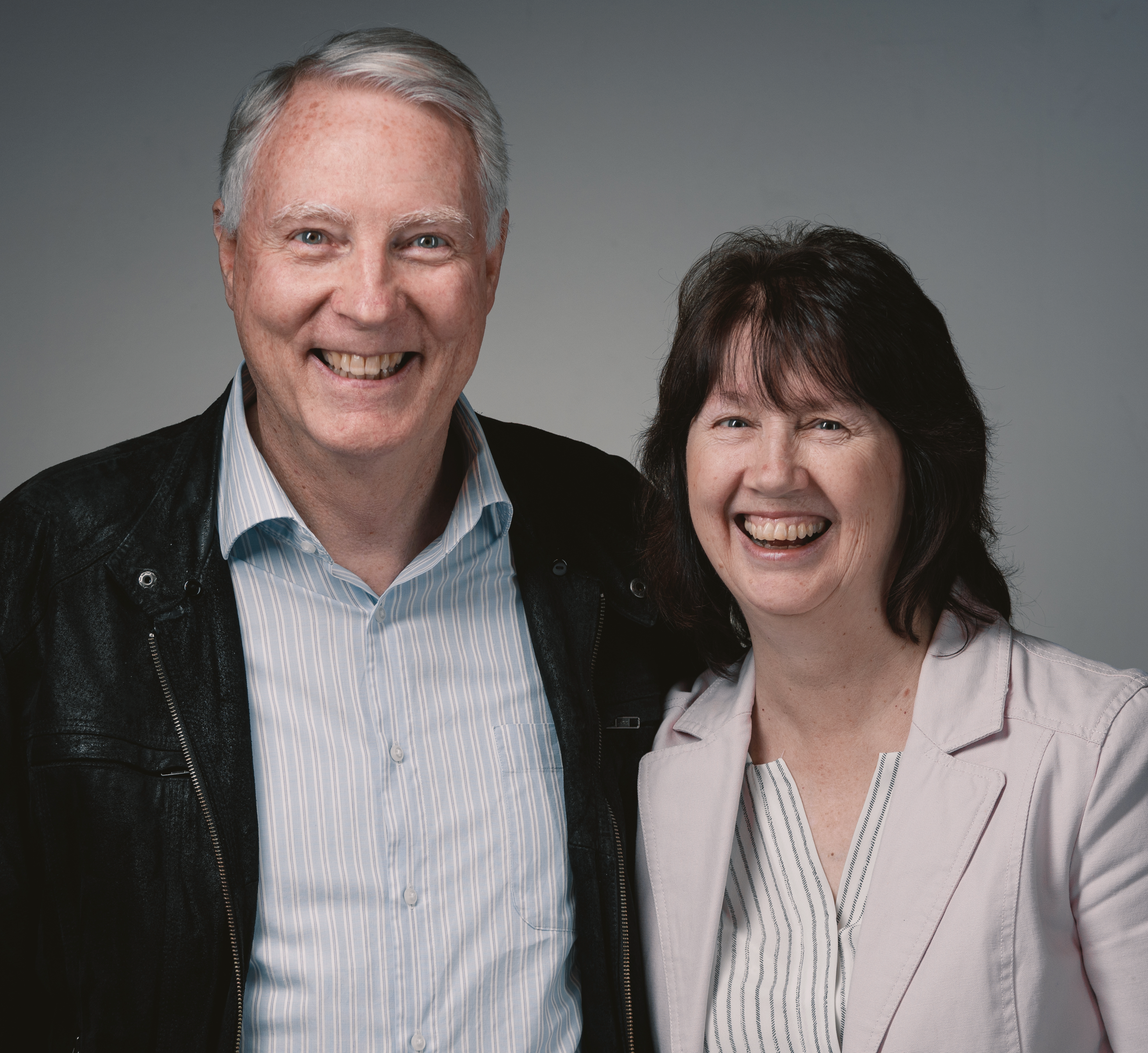 Close-up of a smiling elderly man with gray hair and a woman with dark hair, both in professional attire, against a gray background.