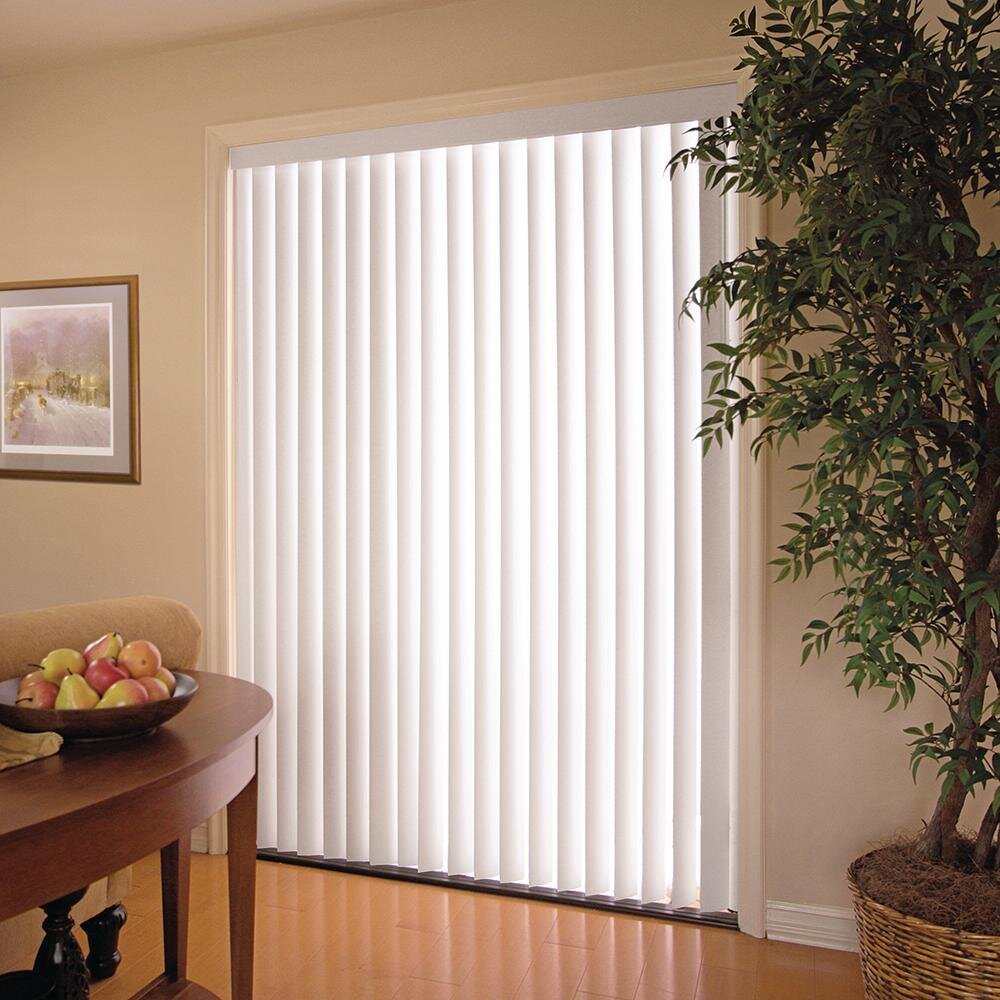 Interior view of a room with vertical white blinds covering a sliding glass door, a large potted plant on the right, a small table with a bowl of apples on the left, and a framed picture on the wall.