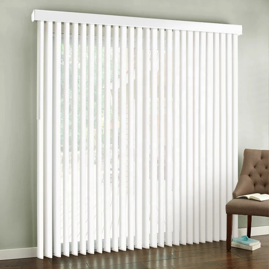 White vertical blinds covering a large window with trees visible outside, sitting in a room with light green walls, a brown tufted chair with an open book on it, and wooden flooring.