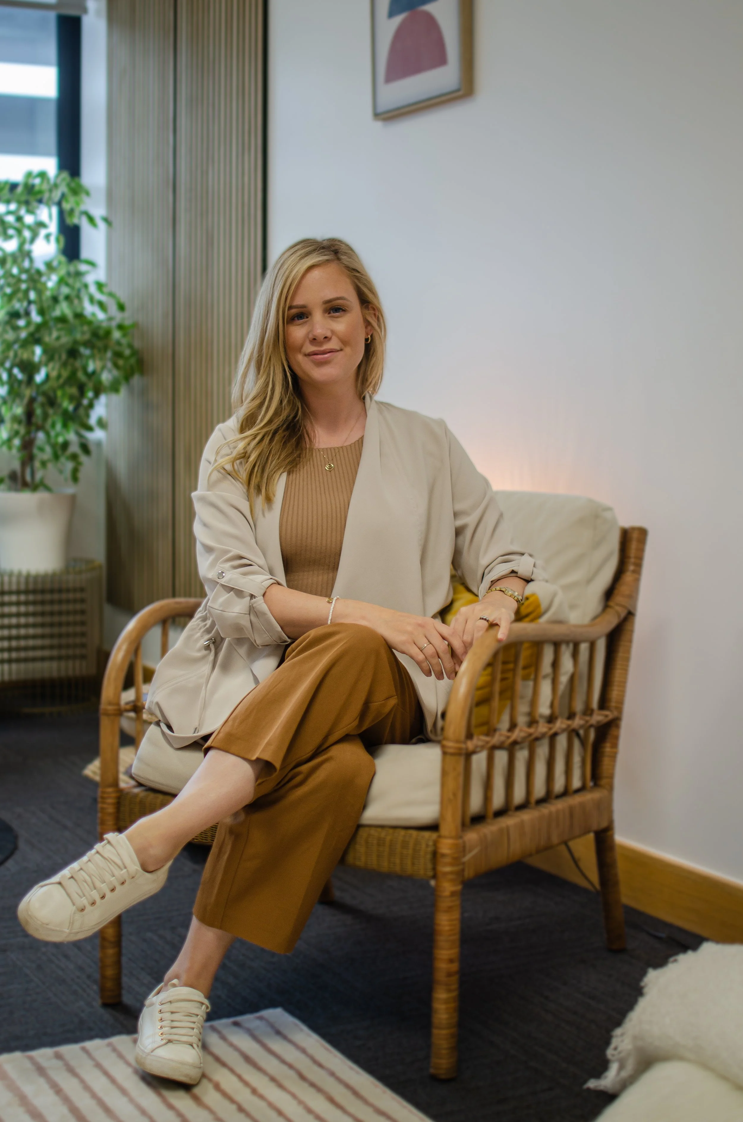 A woman sitting on a wicker chair in a modern, well-lit indoor space, with a plant and framed art on the wall behind her.