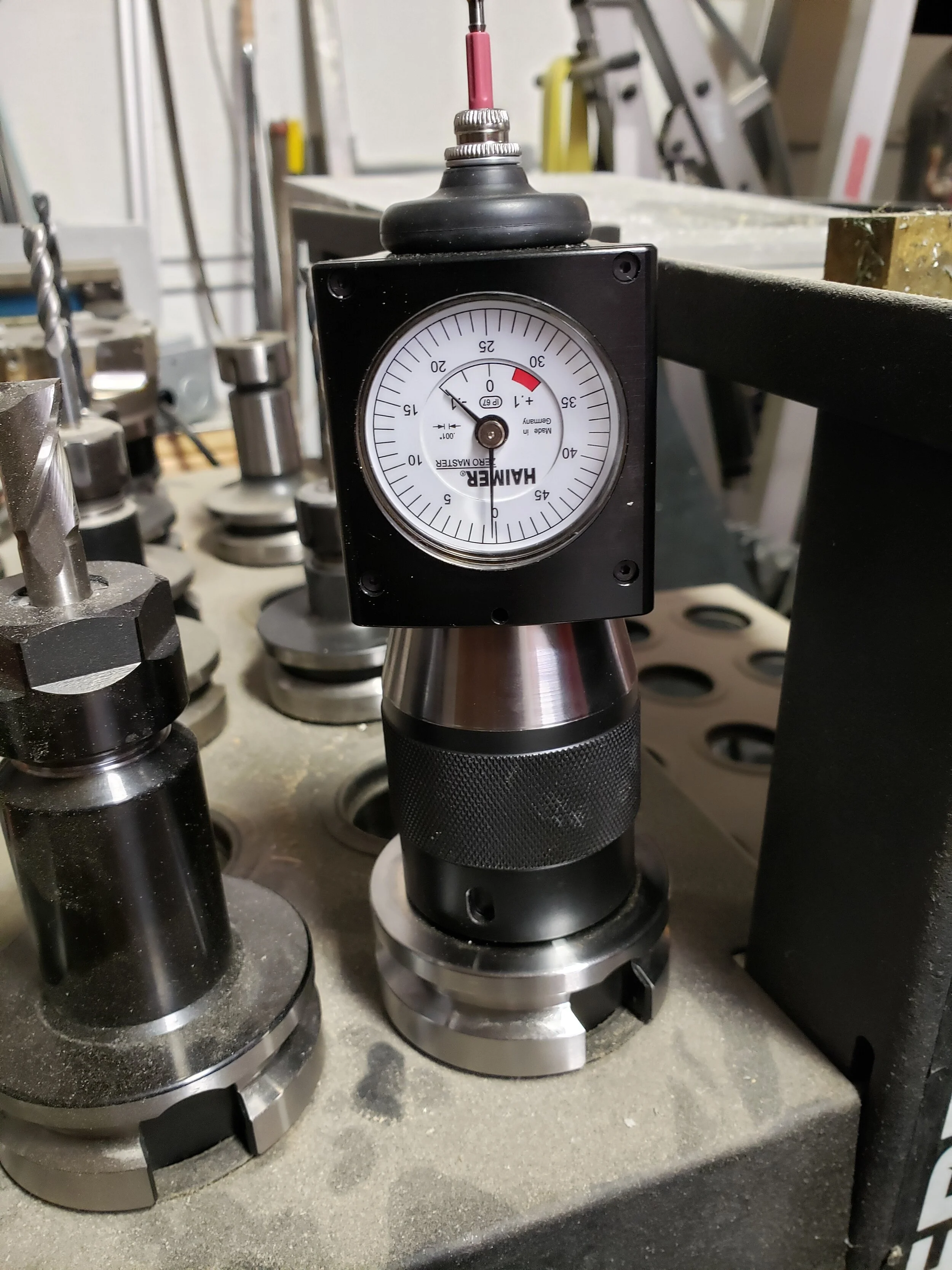 A dial indicator gauge mounted on a machine, surrounded by various drill bits and metalworking tools in a workshop or manufacturing setting.
