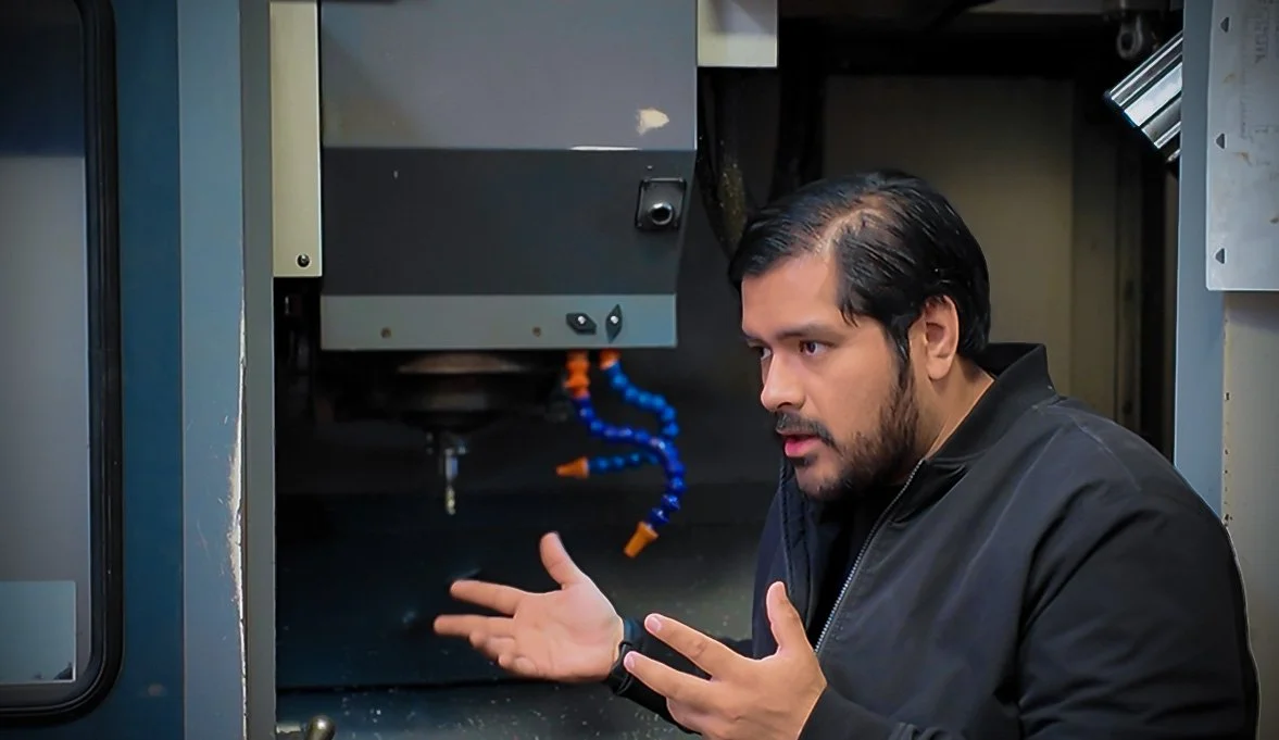 Man explaining inside a machine shop, with a CNC machine in the background.
