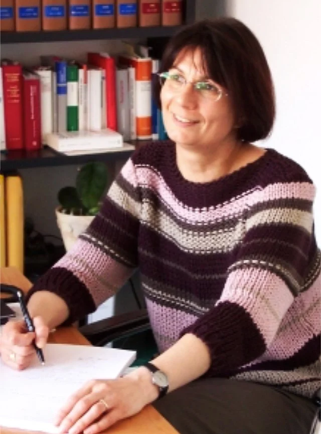 Frau mit dunkelbraunen Haaren und Brille, sitzt an einem Tisch mit Papier und Stift, im Hintergrund Bücherregal mit bunten Büchern.