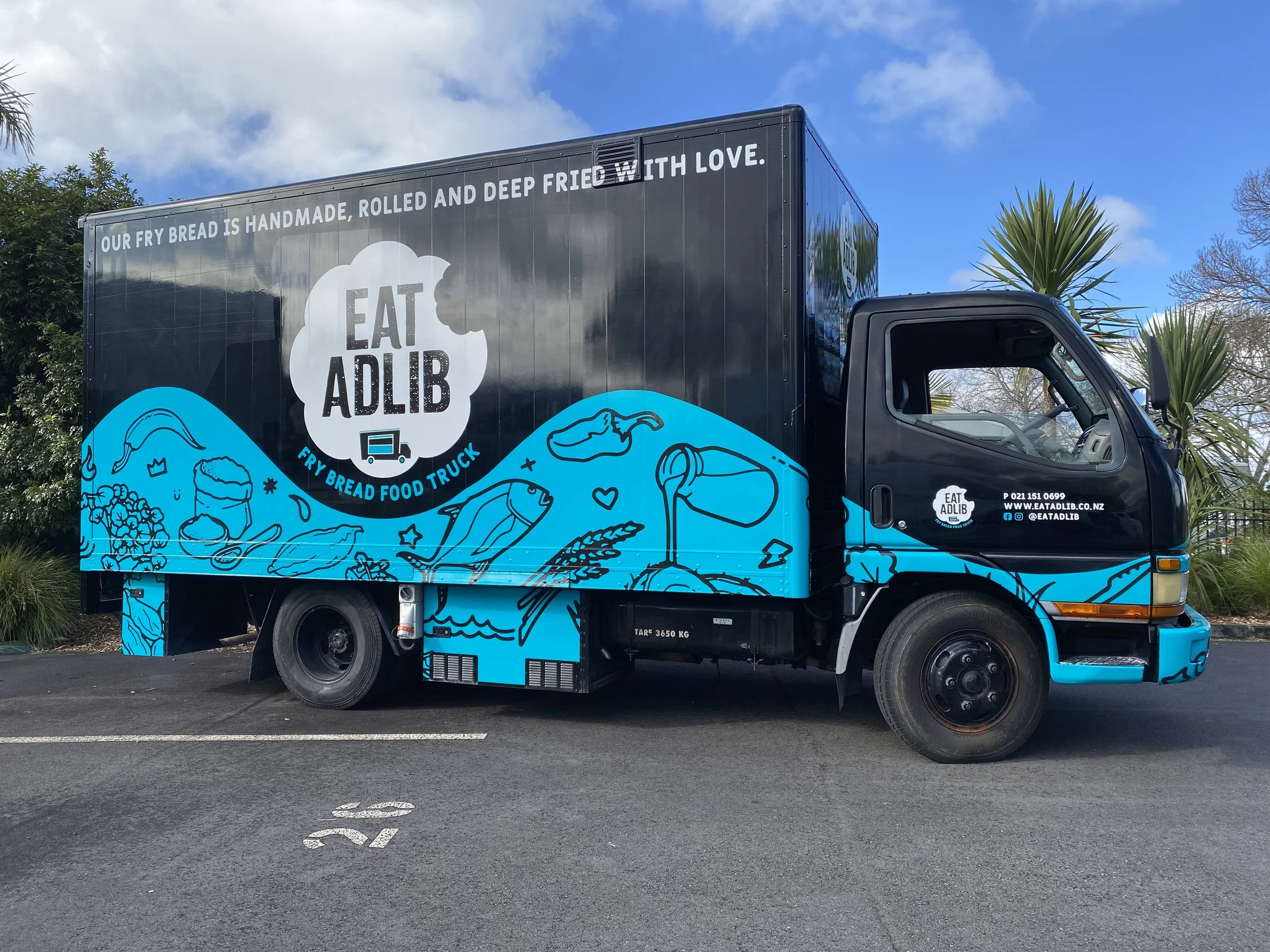 Food truck with "Eat Adlib" branding and fry bread theme, featuring illustrations and contact information. Parked outdoors with clear sky in background.