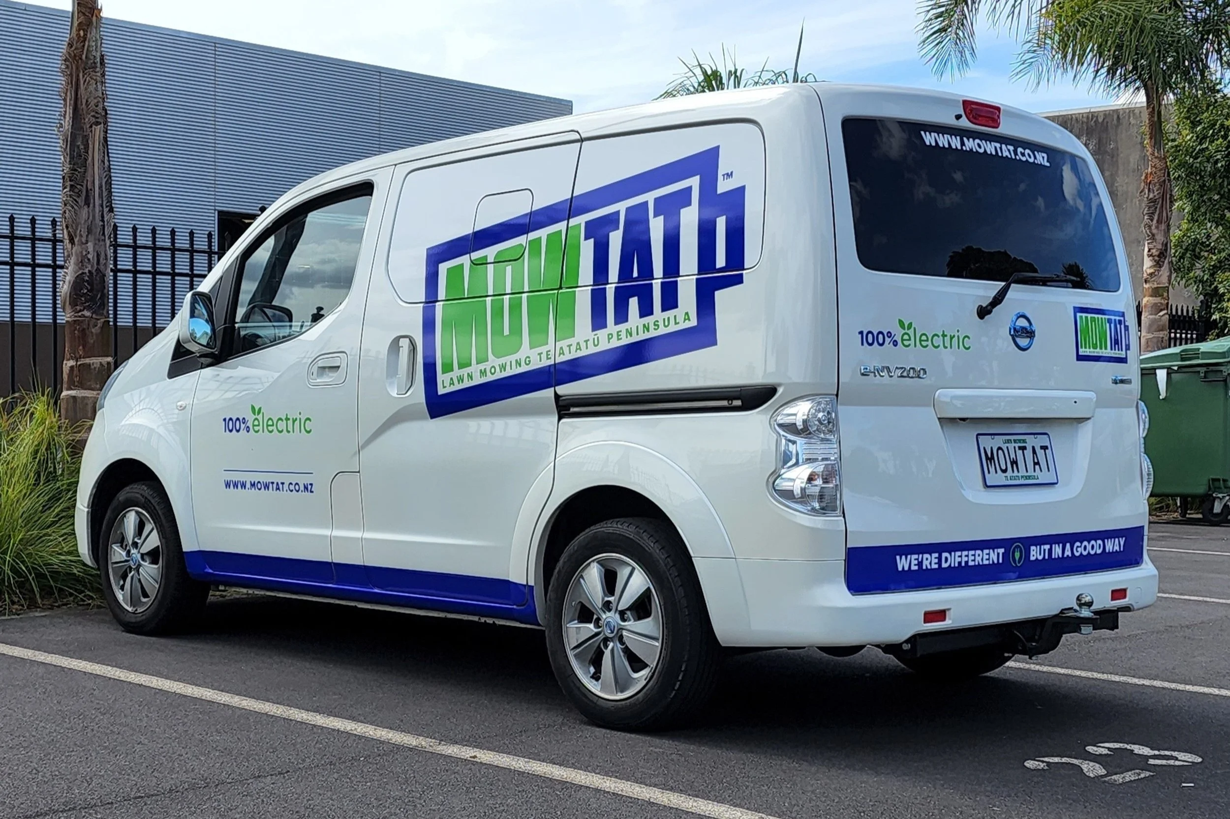 A white electric van in a parking lot featuring MowTat advertising, with green '100% electric' text and a blue and green logo. The license plate reads 'MOWTAT'. The van promotes lawn mowing services and has 'We're different but in a good way' slogan.