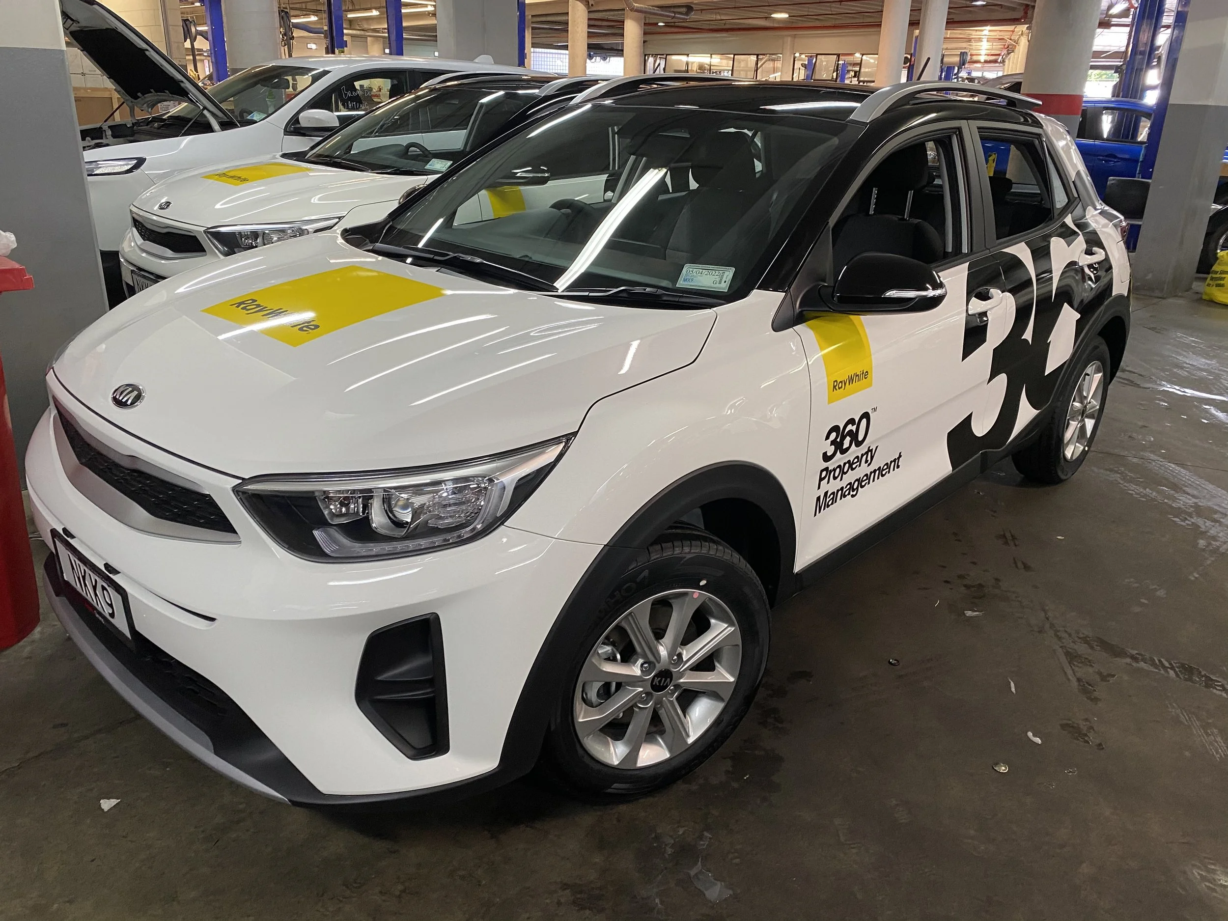 White Kia car with "360 Property Management" branding, parked indoors with other cars.