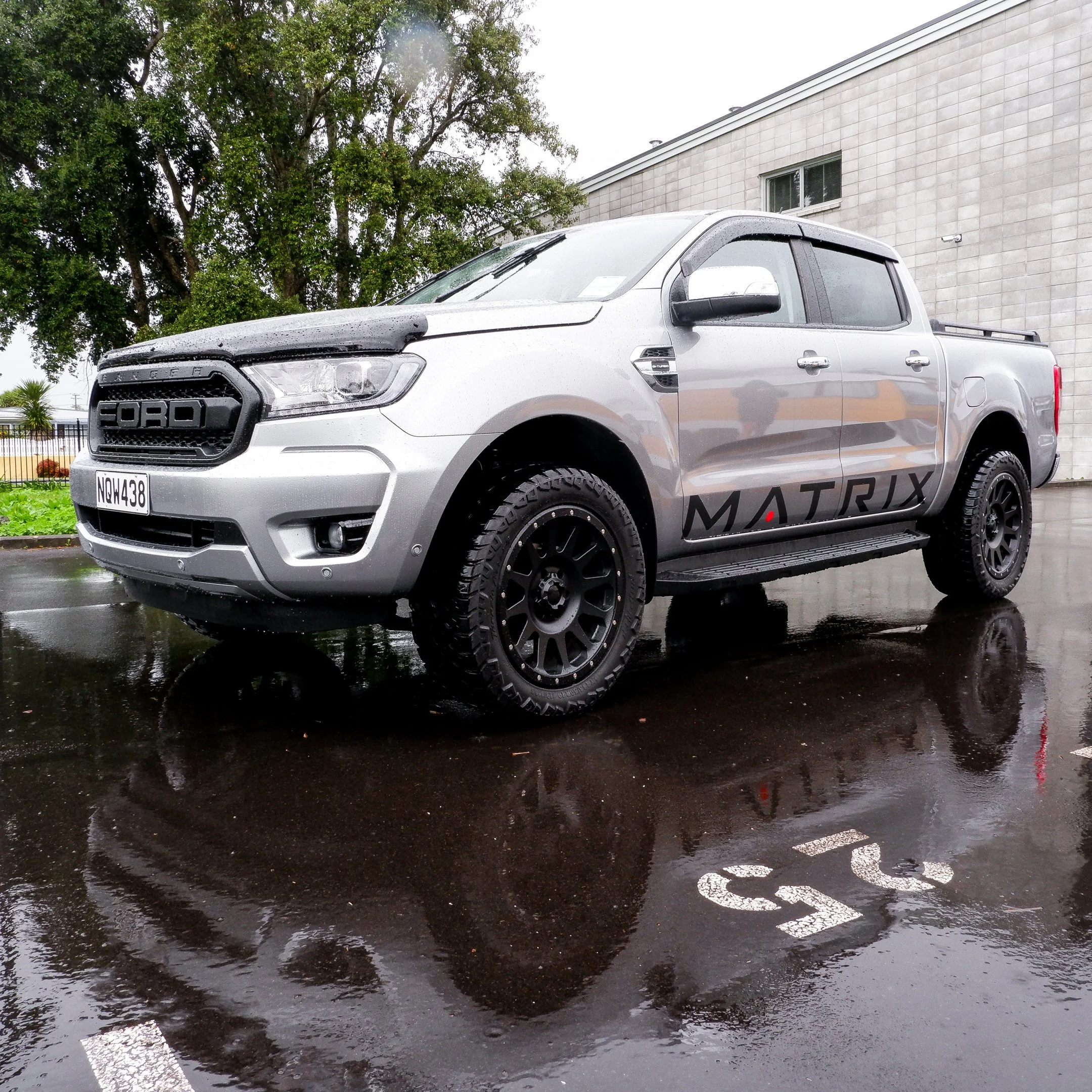 Silver Ford pickup truck with large tires parked on wet pavement, reflection visible, with the word 'MATRIX' on the side, background of trees and a building wall.