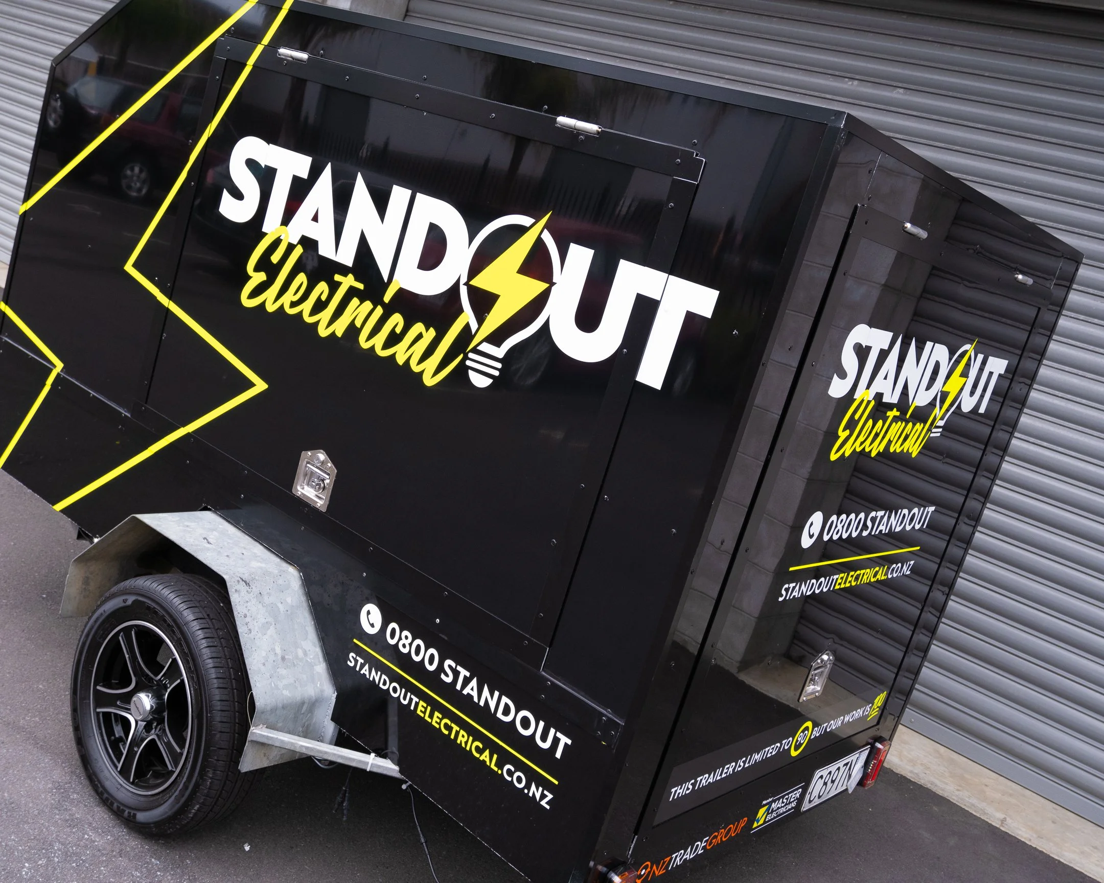 A black trailer labeled 'Standout Electrical' with a yellow lightning bolt and lightbulb logo parked in front of a metal garage door. The trailer displays contact information including a phone number and website.