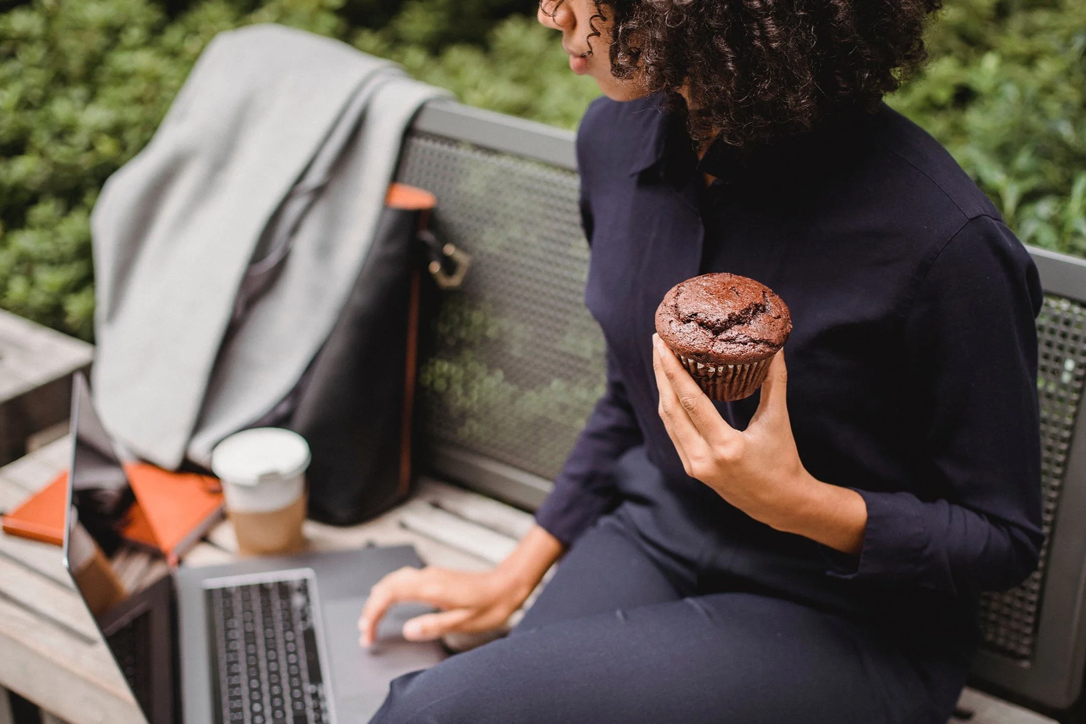 A woman sitting on a park bench holding a chocolate muffin in her right hand, with a laptop, coffee cup, and a backpack nearby.