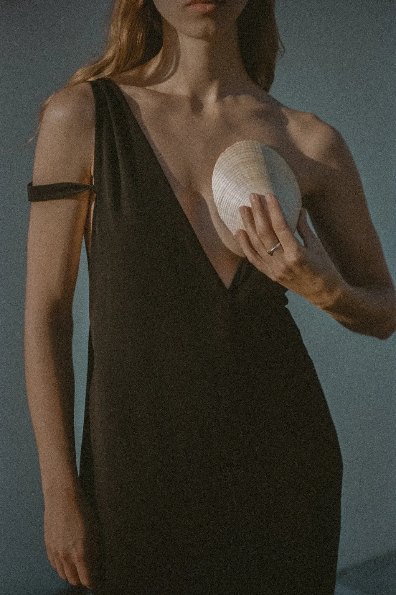 A woman wearing a black dress with a shoulder strap is holding a white seashell close to her chest.