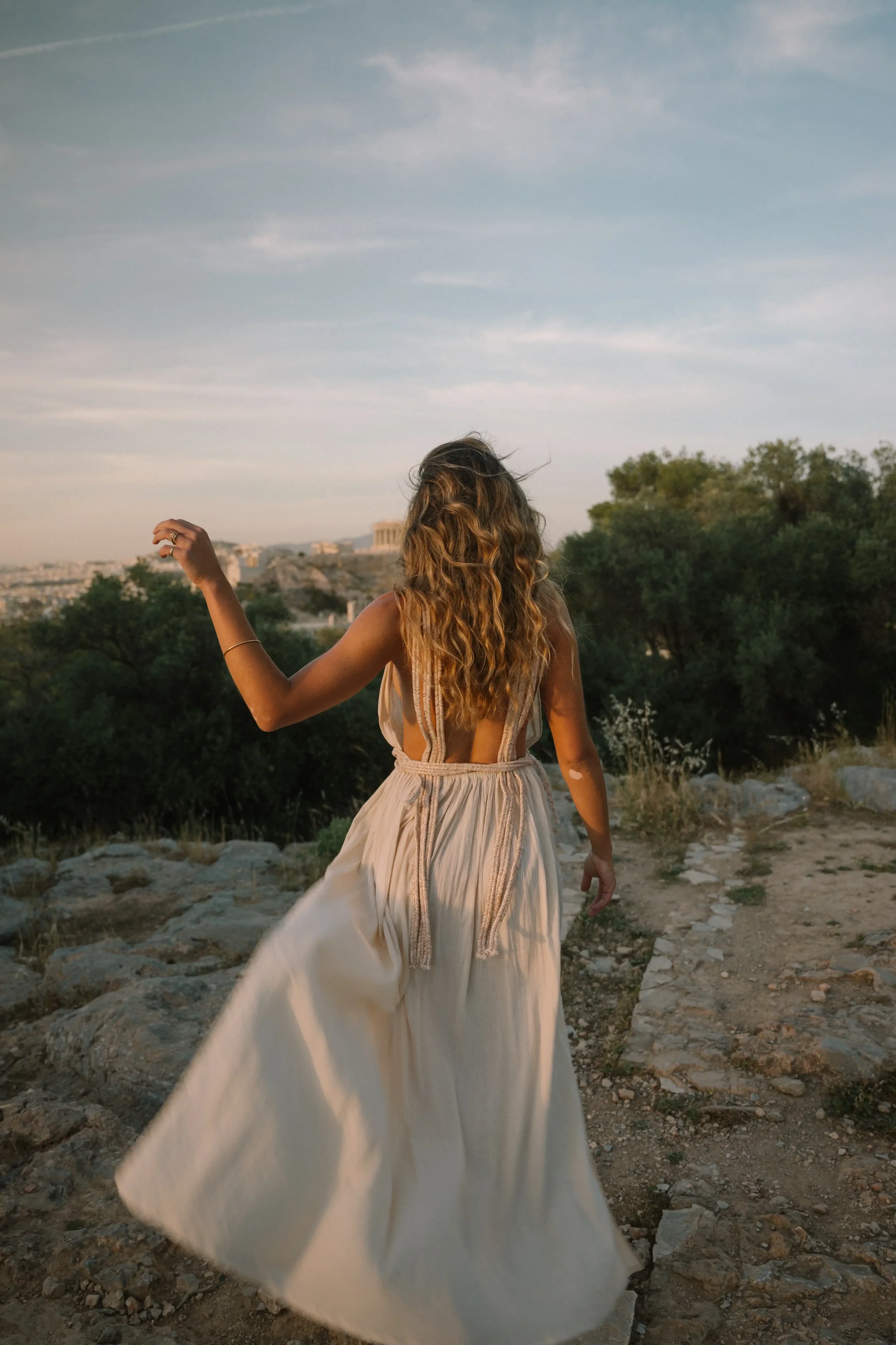 A woman with long wavy hair in a flowing beige dress walking on rocky terrain with a cityscape and ancient ruins in the background during sunset.