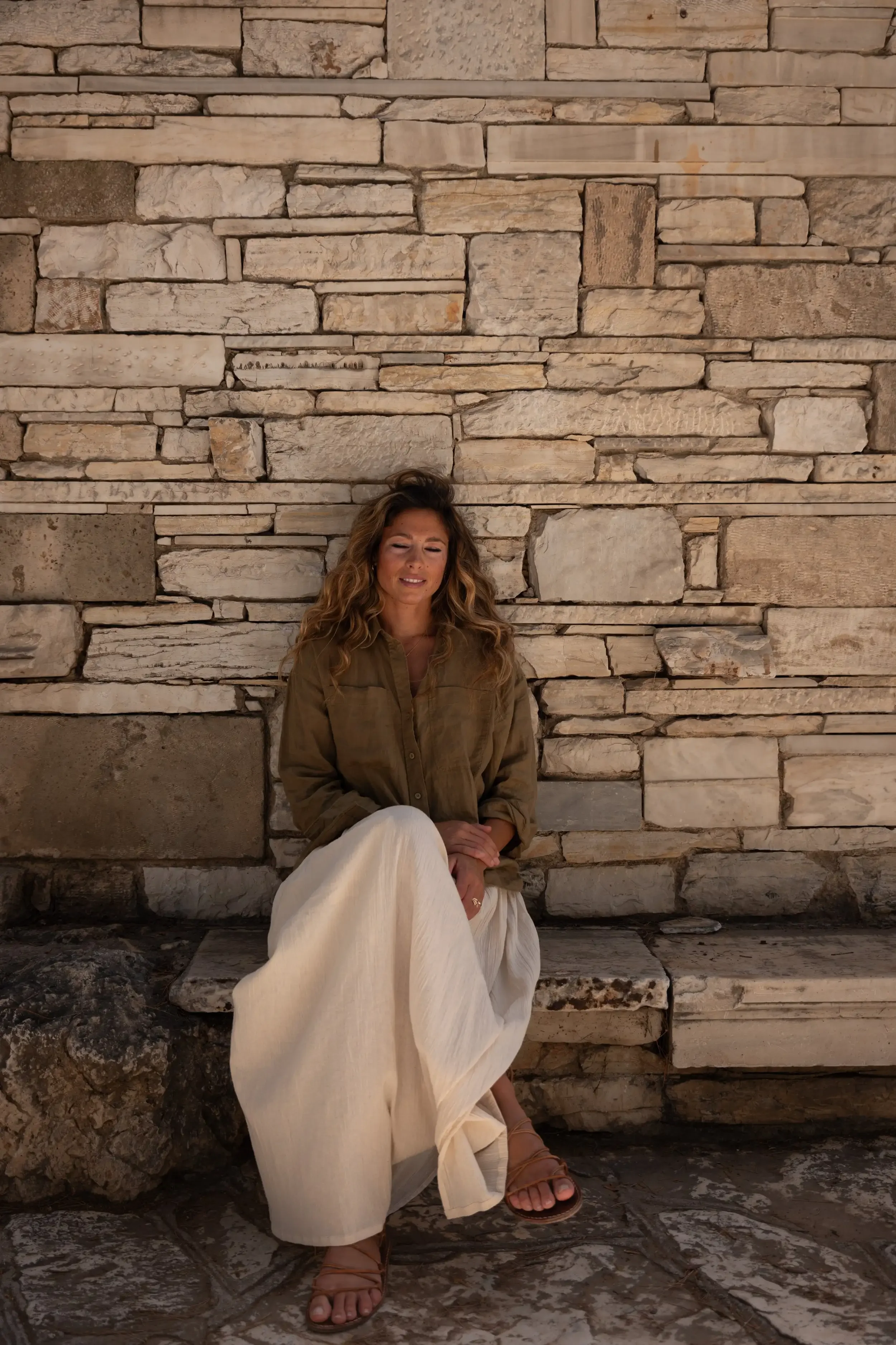 A woman with long, wavy hair sits against a large stone wall, smiling with her eyes closed. She wears a brown jacket, white wide-leg pants, and brown sandals.