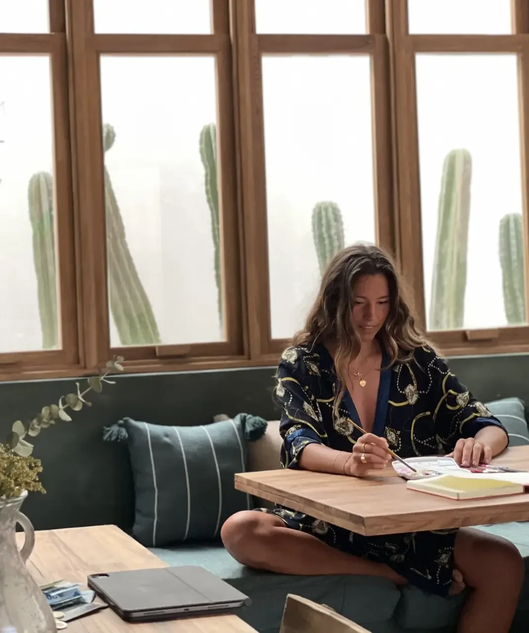 Woman with wavy hair sitting cross-legged at a wooden table, writing in a notebook, with a laptop and flower vase nearby, in front of large windows with cactus plants outside.
