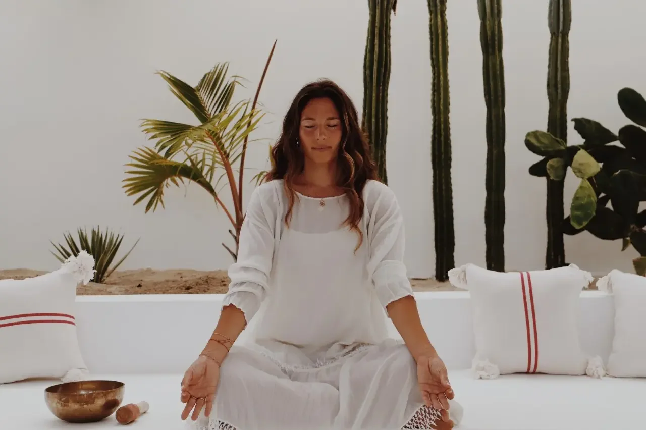 Woman practicing meditation in a bright white room decorated with tropical plants. She is wearing white and sitting cross-legged with her eyes closed.