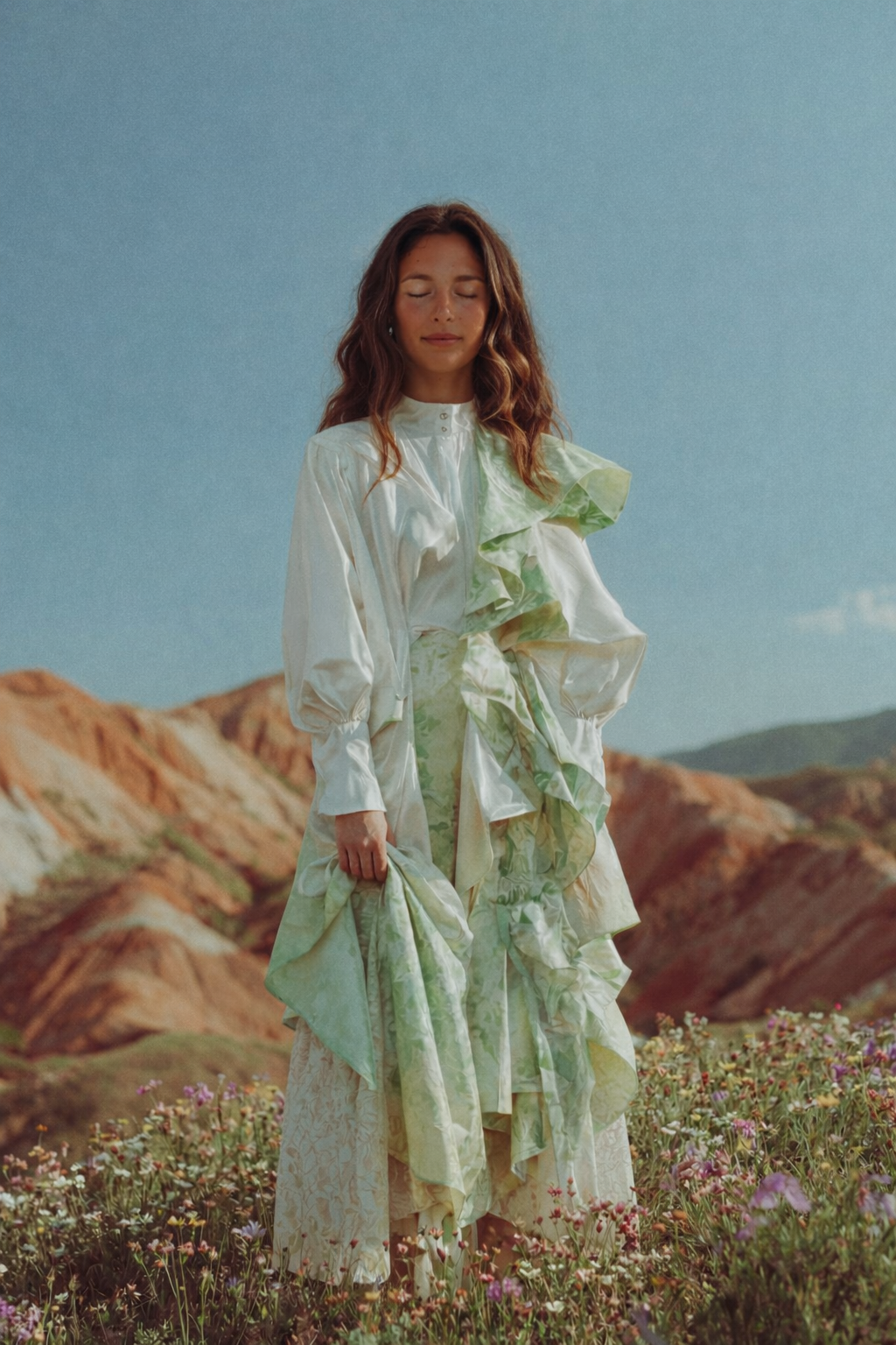 A woman standing in a field of wildflowers with colorful mountains in the background, wearing a white and green ruffled dress with long sleeves.