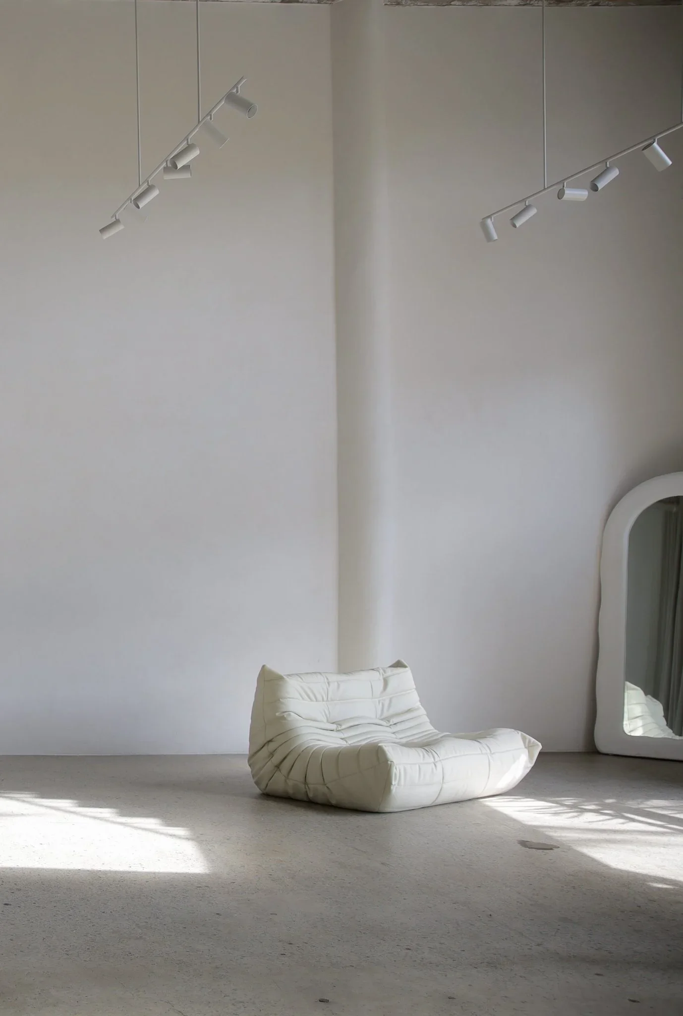 Minimal white interior with a low cushioned chair, soft daylight, and a standing mirror.