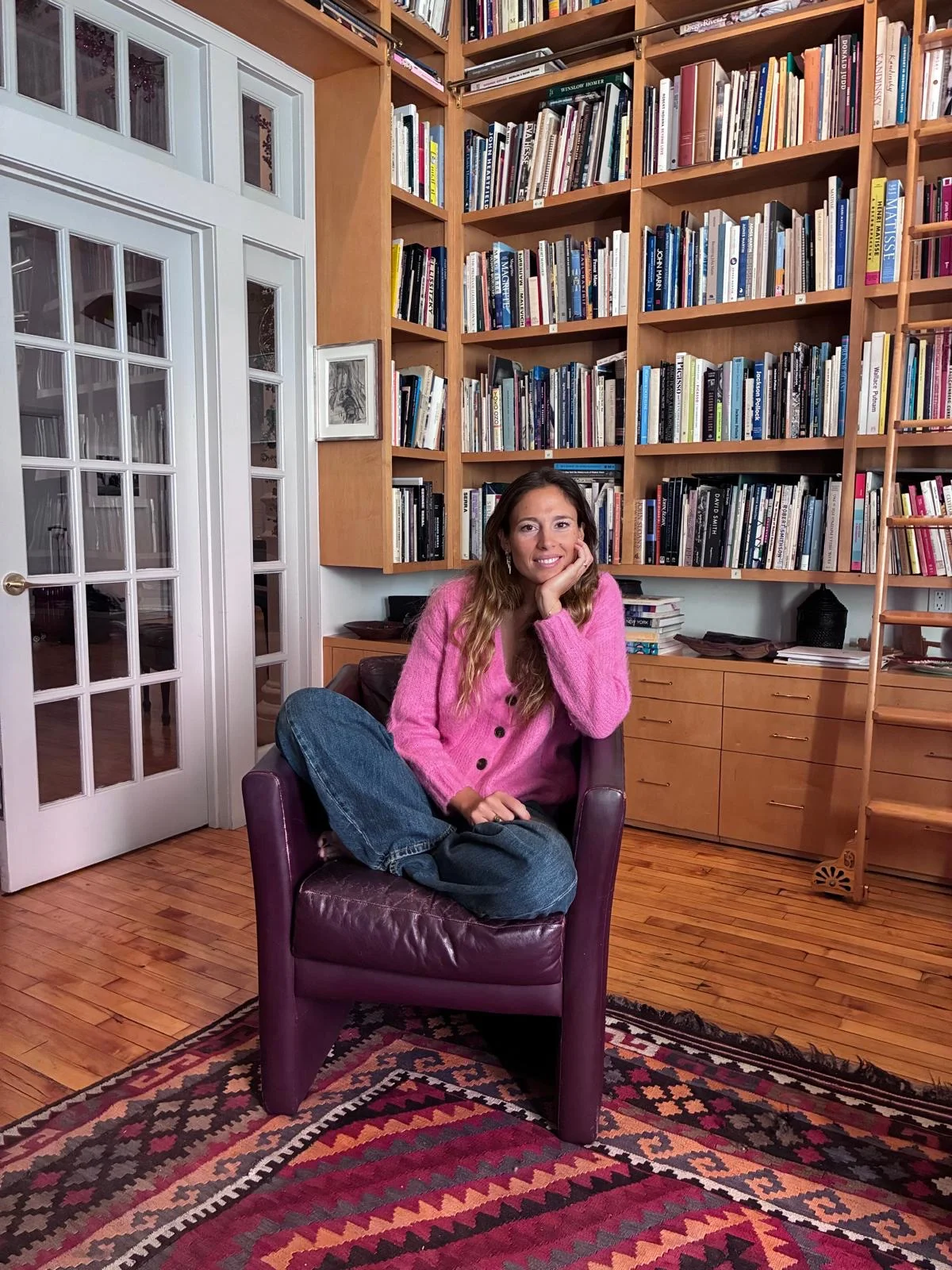 A woman sitting in a purple armchair in a room with wooden floors and a large bookshelf filled with books. She is wearing a pink sweater and blue jeans, smiling with her hand resting on her cheek.