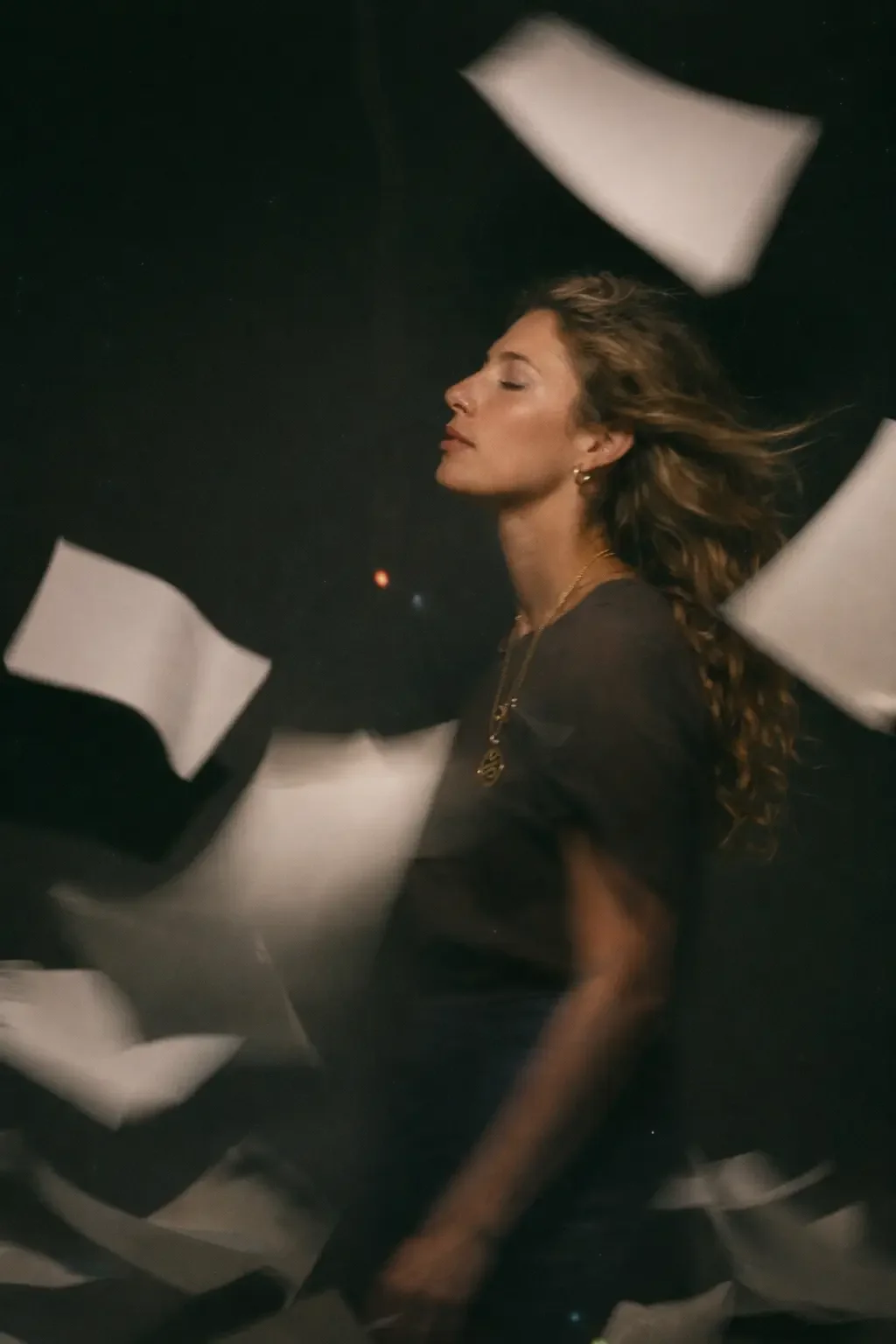 A woman with long curly hair and gold jewelry has her eyes closed and head slightly tilted back against a dark background, with white papers floating around her.