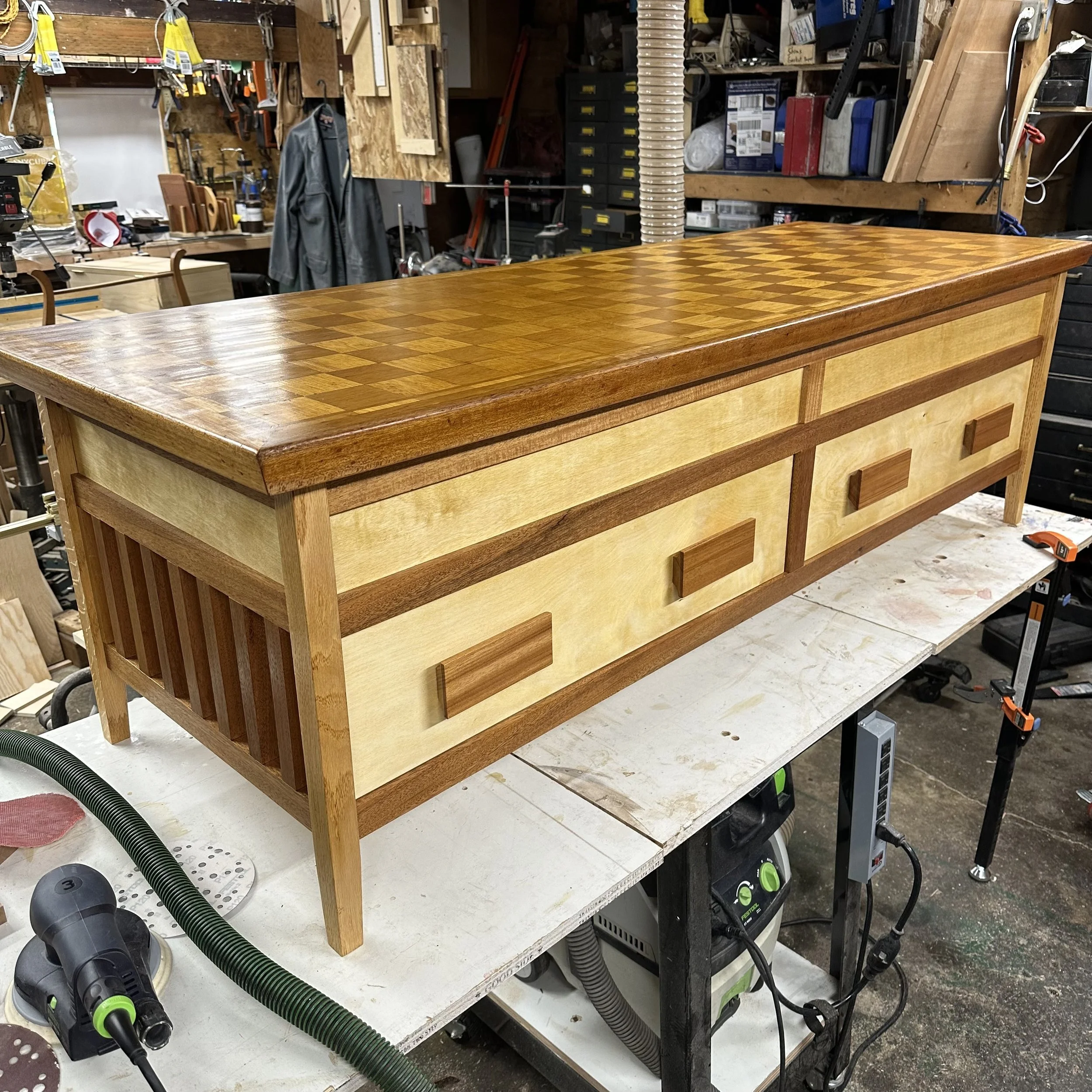 Custom wooden sideboard with a checkered top surface, storage drawers, and slatted sides in a woodworking workshop.