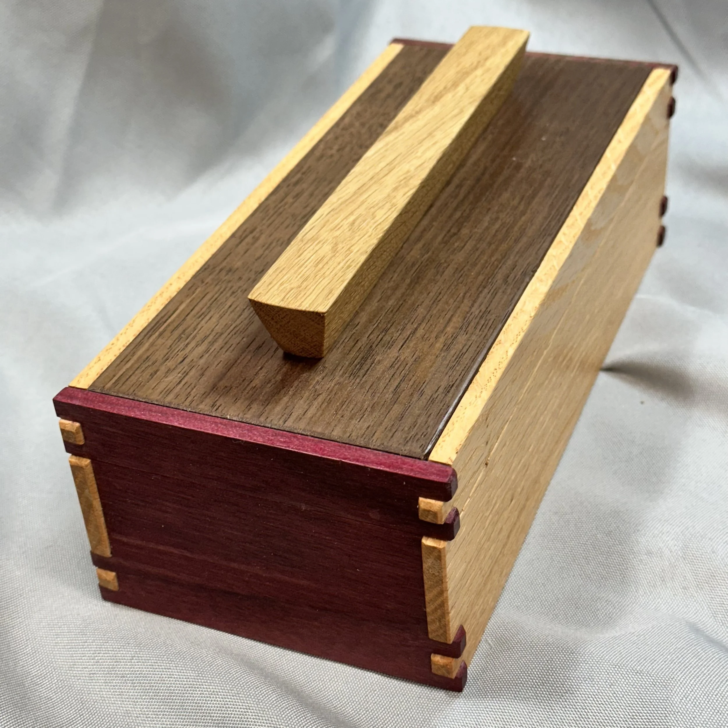 A small wooden box with a handle on top, made of different colored wood pieces including dark brown, light brown, and maroon, with visible box joints at the corners.