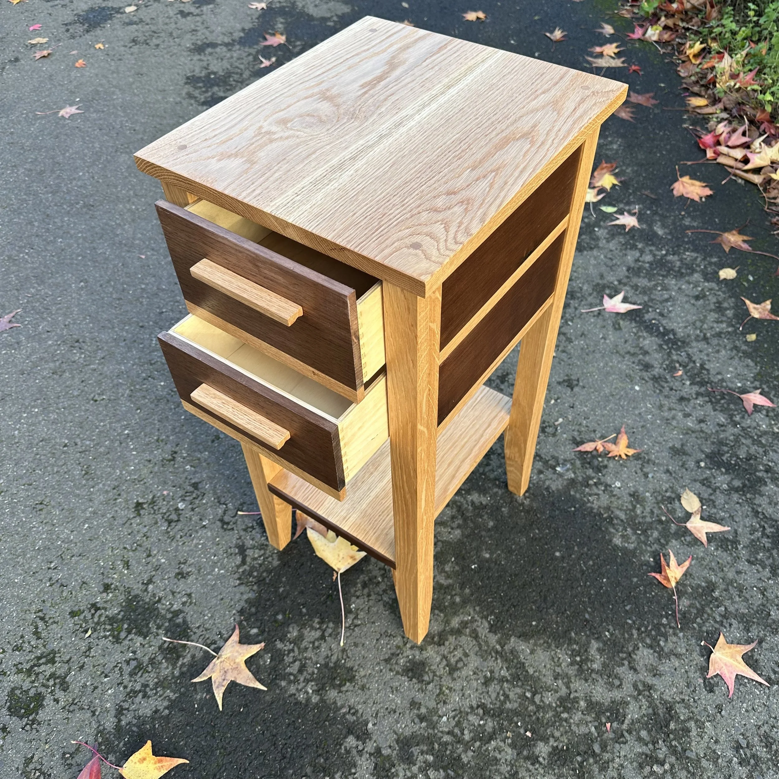 A wooden nightstand with two open drawers, placed outdoors on a paved surface with fallen autumn leaves.
