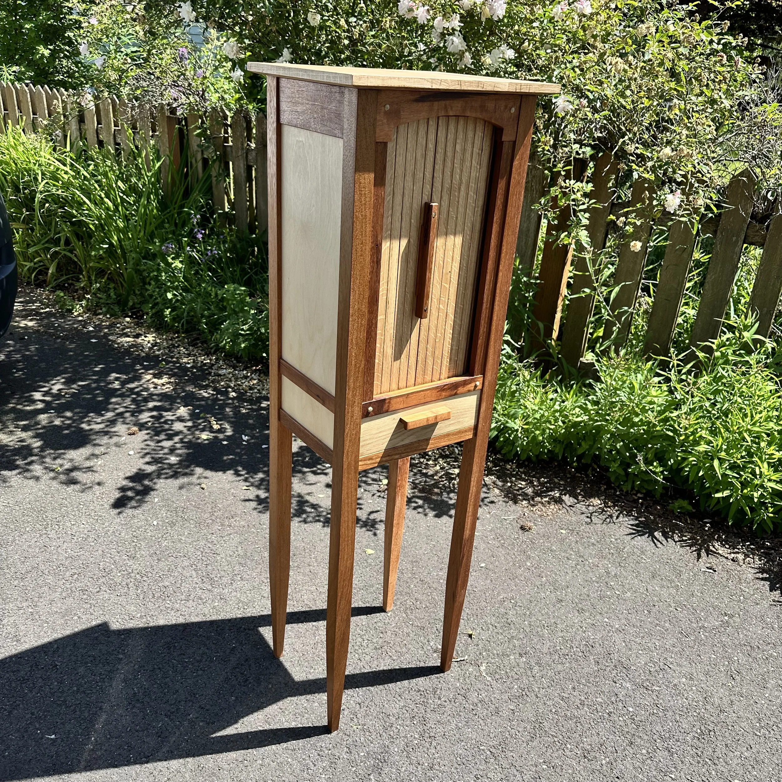 Wooden whiskey cabinet with a tambour door, drawer, and tall legs situated on a paved surface with greenery and a wooden fence in the background.