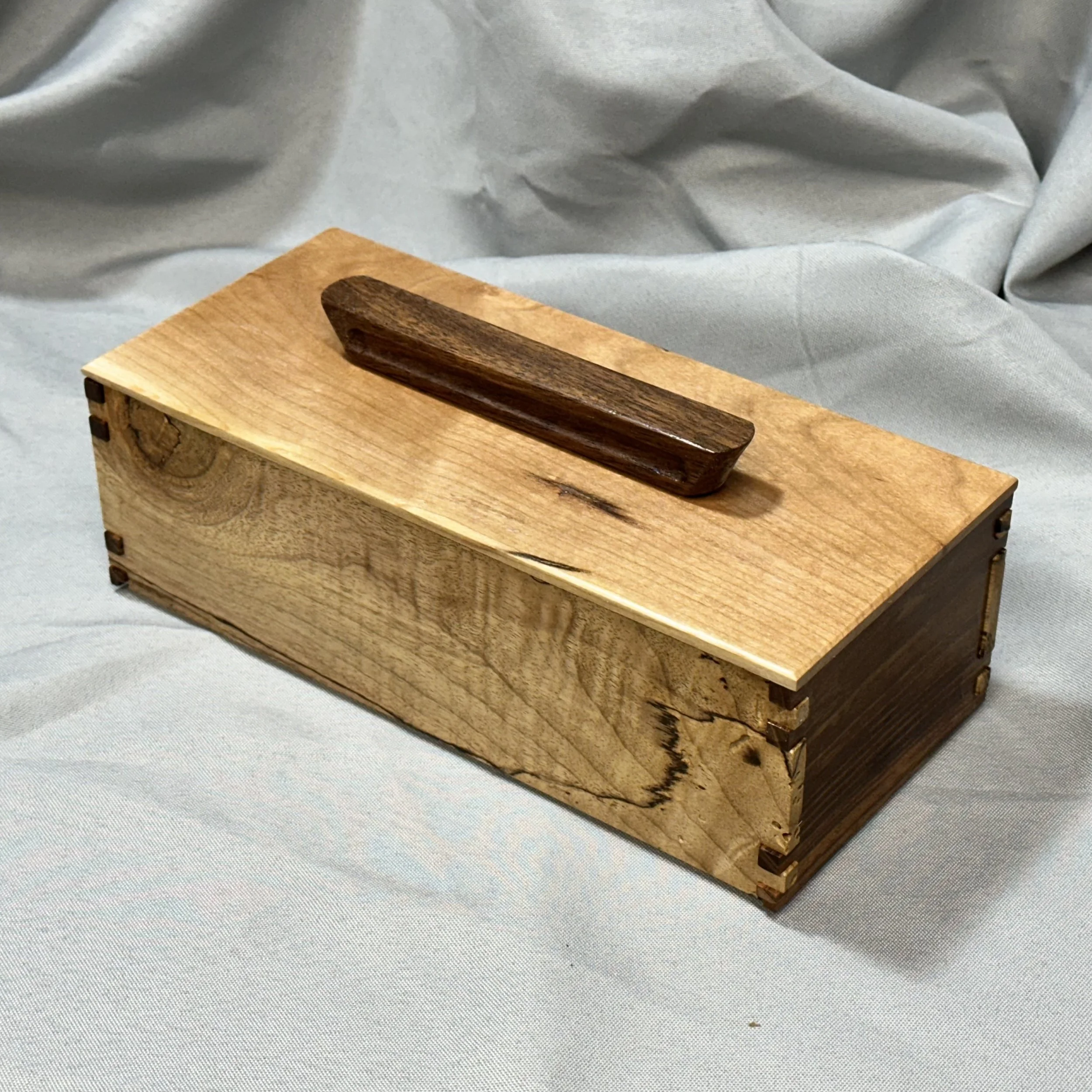 A rectangular wooden box with dovetail joints and a dark wooden handle on top, placed on a light gray fabric background.