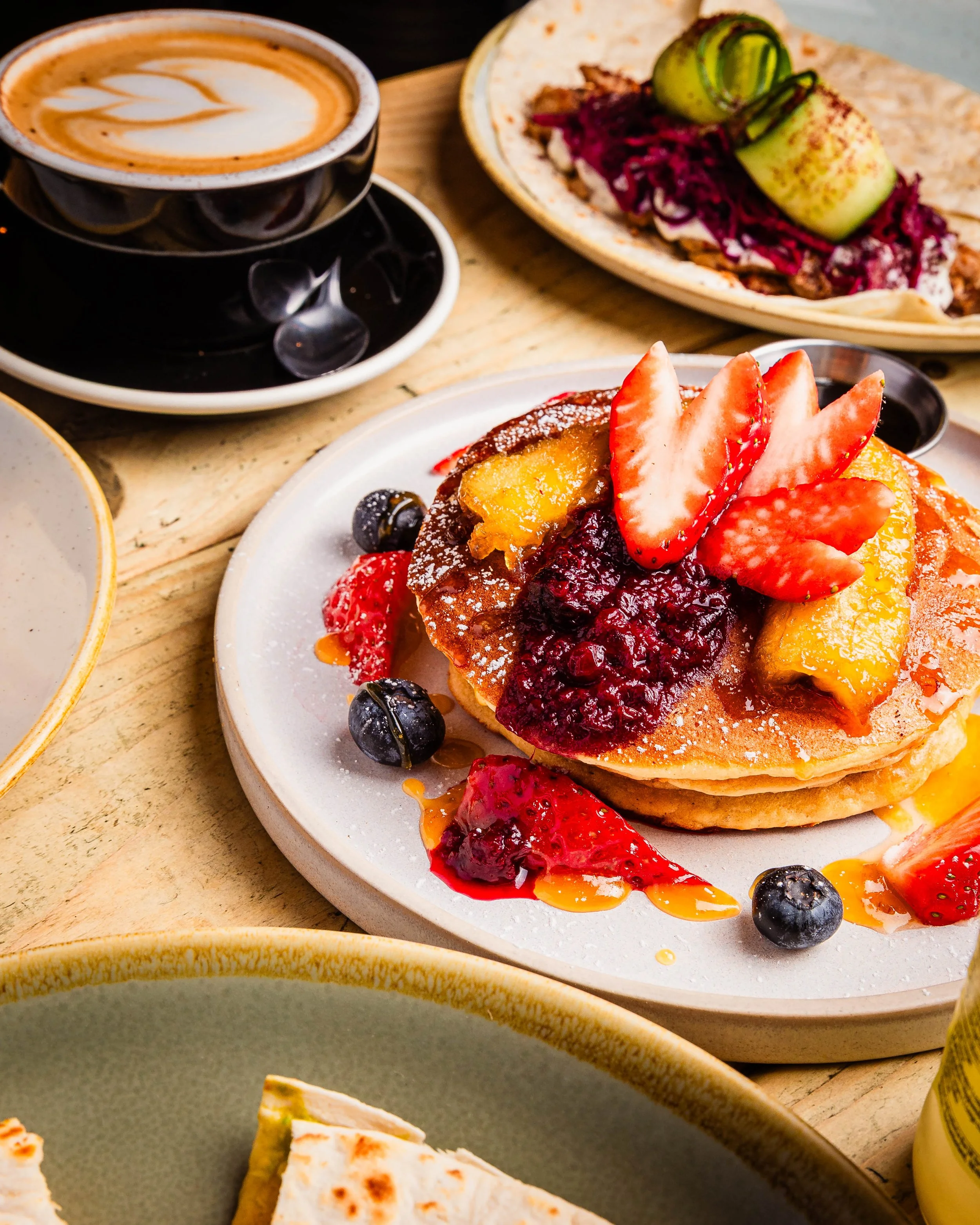 Plate of pancakes topped with strawberries, blueberries, peach slices, and berry compote, with syrup drizzled on top, and a cup of latte with latte art. Perfect brunch in Earlsfield, London.