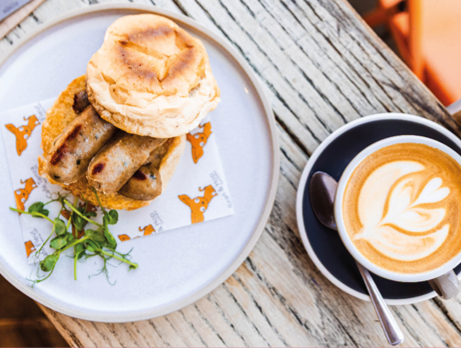 Plate with breakfast items including toasted bread and sausage rolls, garnished with microgreens, alongside a cup of latte with latte art on a wooden table. A welcoming local café where regulars feel at home